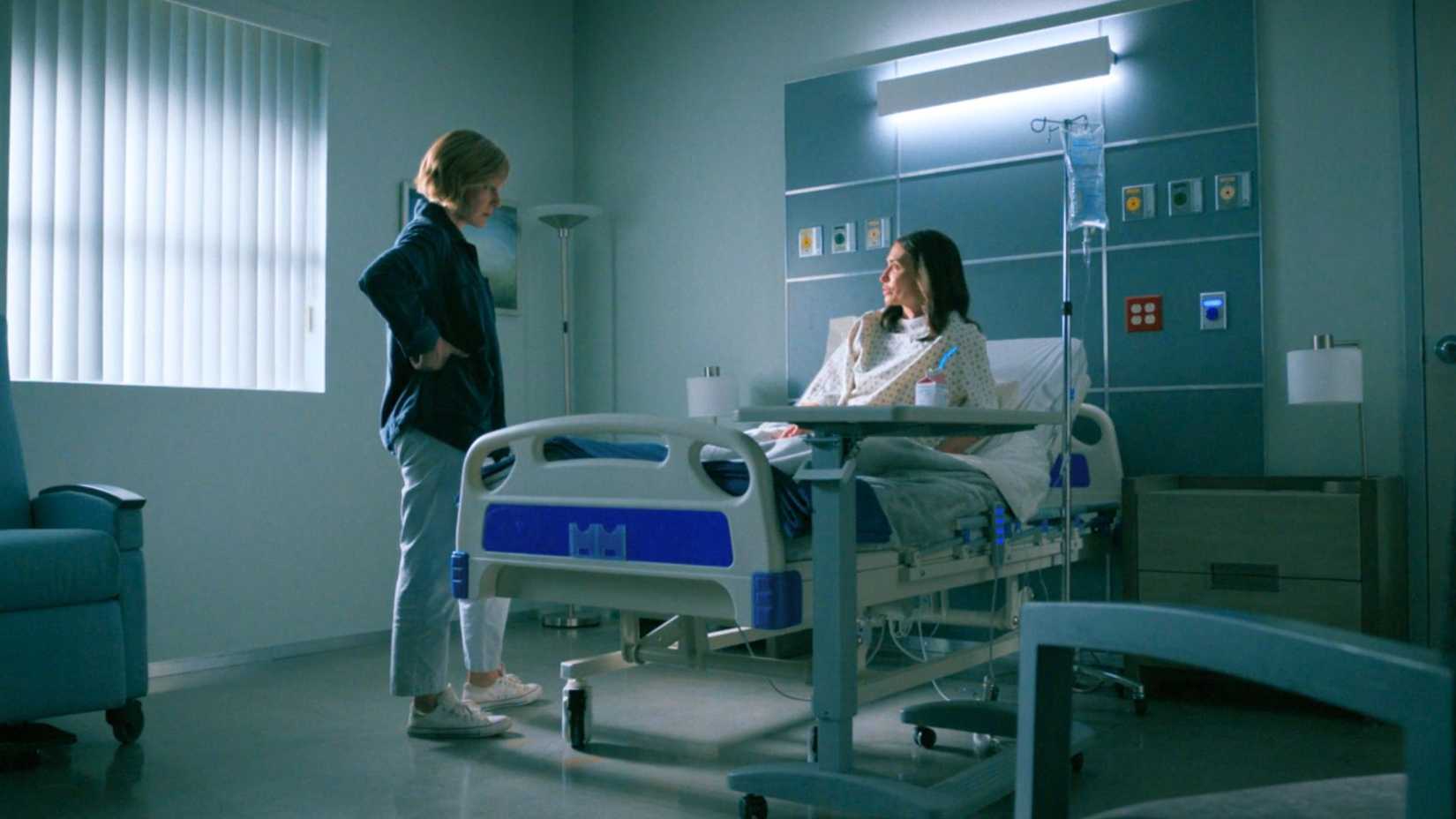 A screenshot from Pluribus episode 4 featuring Rhea Seehorn as Carol Sturka and Karolina Wydra as Zosia. Carol is visiting Zosia, who lies in a hospital bed.