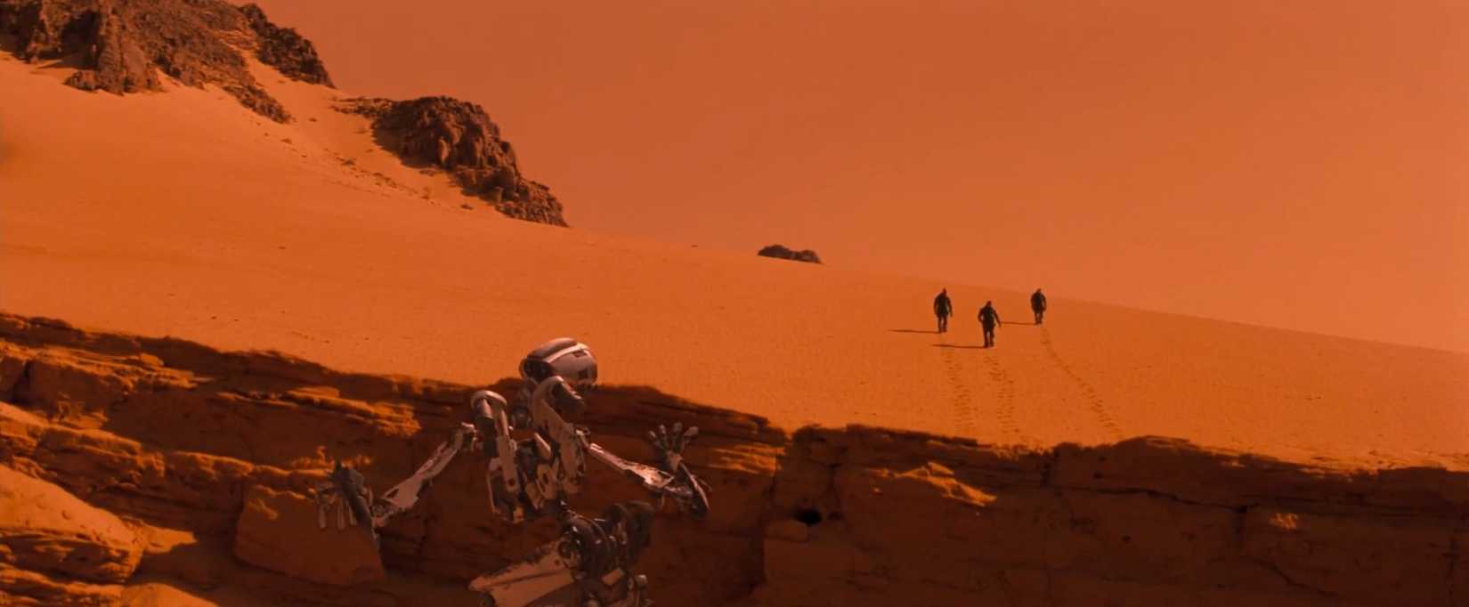 A scene from the film Red Planet depicts the vast Martian landscape in a red-orange wide shot, with the three astronauts dwarfed in the distance. 