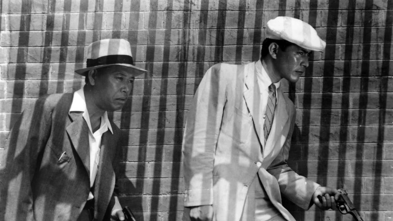 Two Japanese men in suits and hats, one brandishing a gun, walk next to a brick wall, bathed in stripes of light and shadow that fall over them like bars, in Akira Kurosawa's Stray Dog