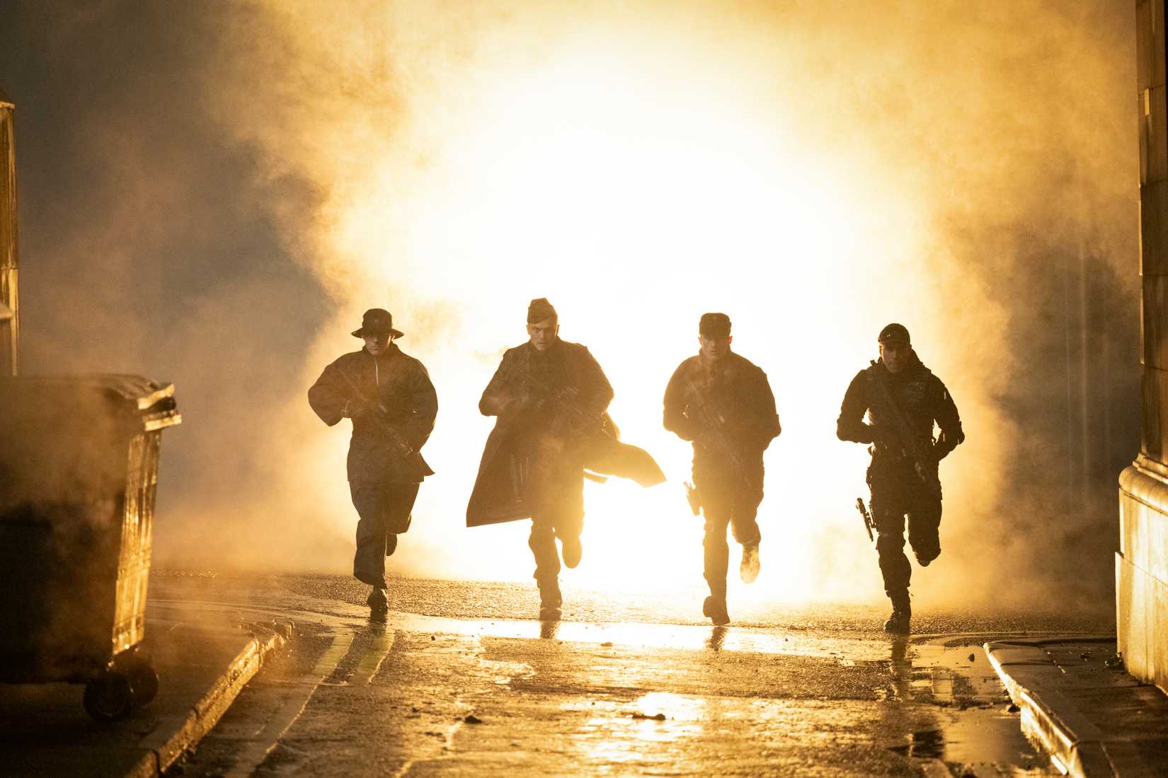 Four heavily armed men in masks and jackets run toward the camera in silhouette, an explosion behind them, in the 2025 Running Man