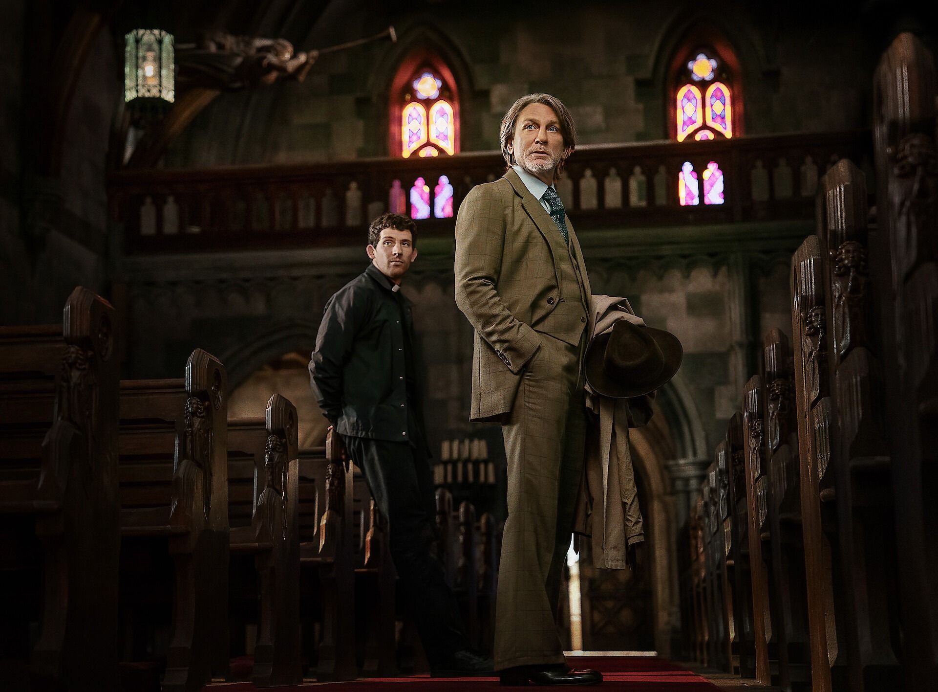 Daniel Craig, as detective Benoit Blanc, and Josh O'Connor, as a priest suspected of murder, look down the aisle of a Catholic church in a scene from Wake Up Dead Man: A Knives Out Mystery.