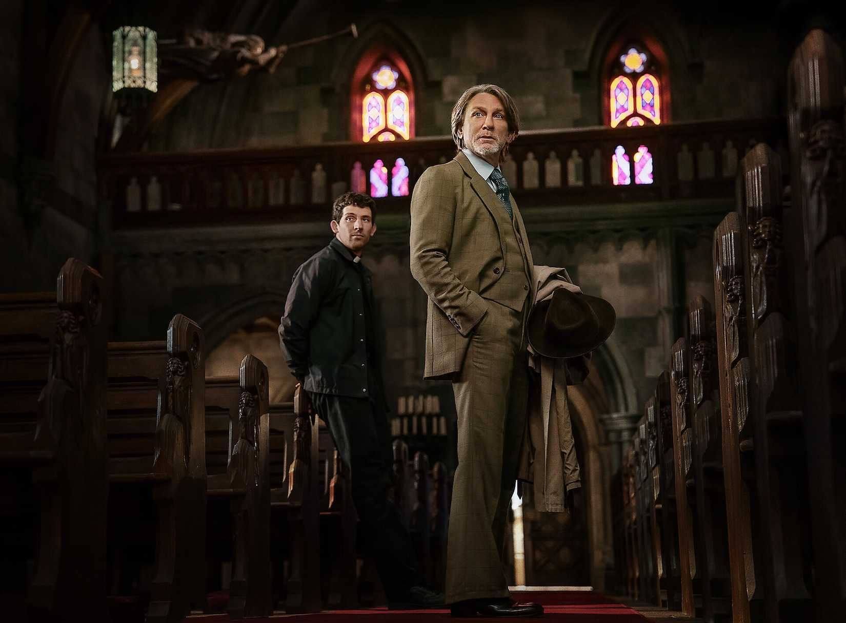 Daniel Craig ในบทนักสืบ Benoit Blanc และ Josh O'Connor ในฐานะนักบวชที่ต้องสงสัยว่าเป็นฆาตกร จ้องมองไปตามทางเดินของโบสถ์คาทอลิกในฉากหนึ่งจาก Wake Up Dead Man: A Knives Out Mystery