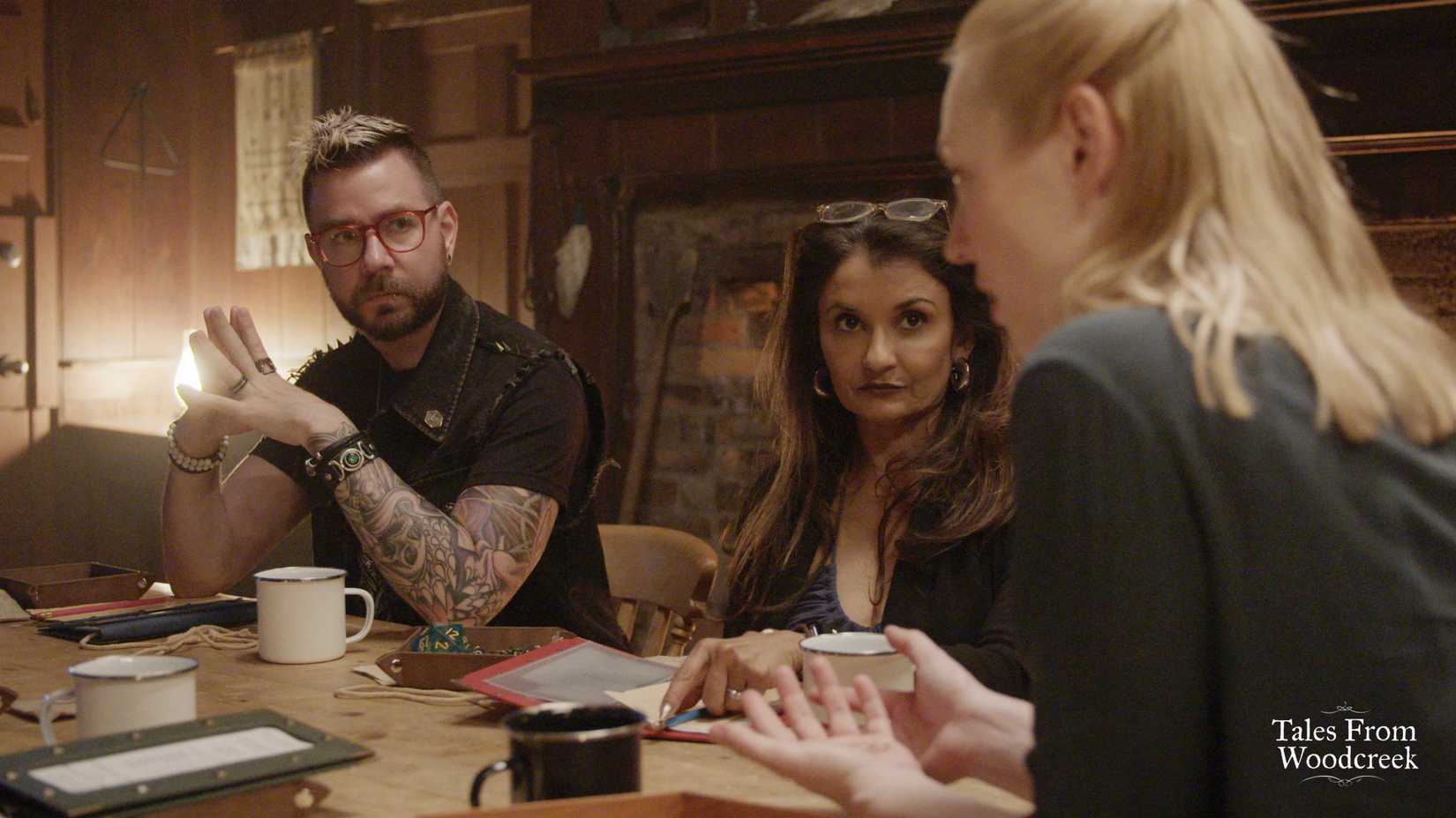 Kelly McLaughlin and Anjali Bhimani sit at a table looking at Dungeon Master Deborah Ann Woll during an episode of Tales From Woodcreek