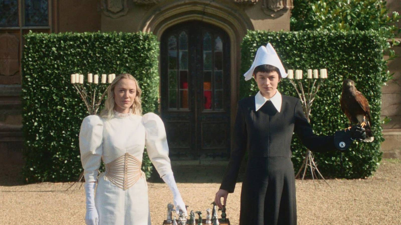 Maika Monroe and Emma Corrin in 100 Nights of Hero; Monroe wears a white chest piece and Corrin a black dress with a white collar, holding a falcon; a chess set sits between them.