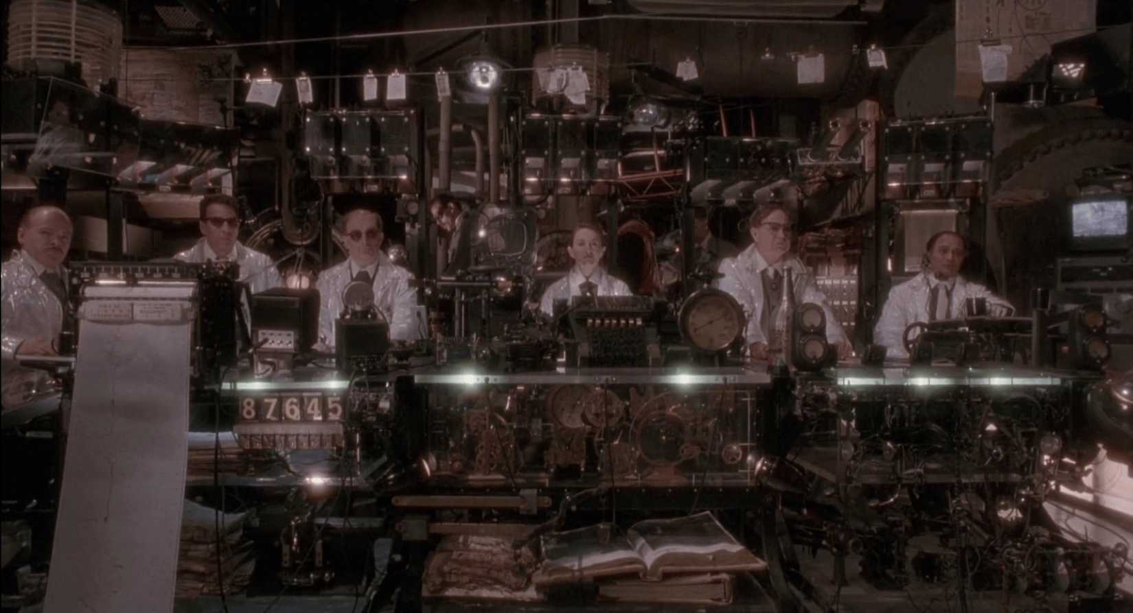 A group of scientists living underground in the cluttered, post-apocalyptic world of 2035 sit in a line interrogating a prisoner (off-screen) in a scene from 12 Monkeys.