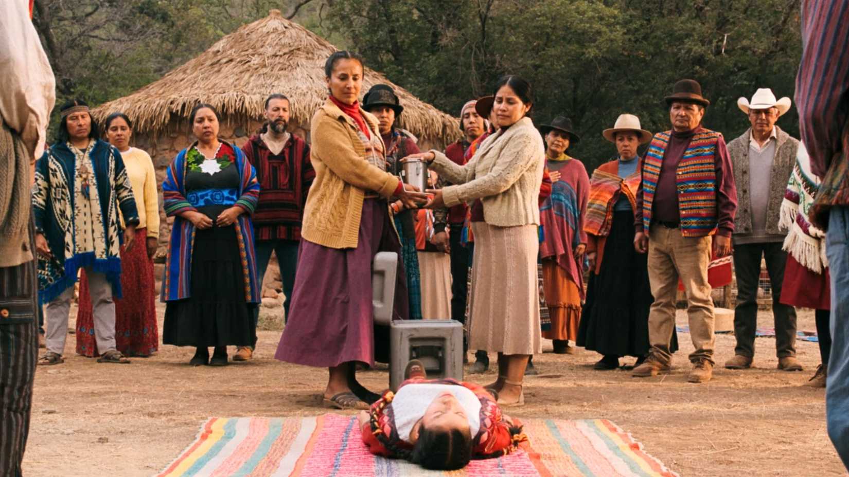 Kusimayu (Darinka Arones) lies on a multicolored blanket. Above her stands two women, holding a metal canister containing the hivemind virus. Surrounding them is the entirety of her village in Peru, all wearing hats and Peruvian attire.