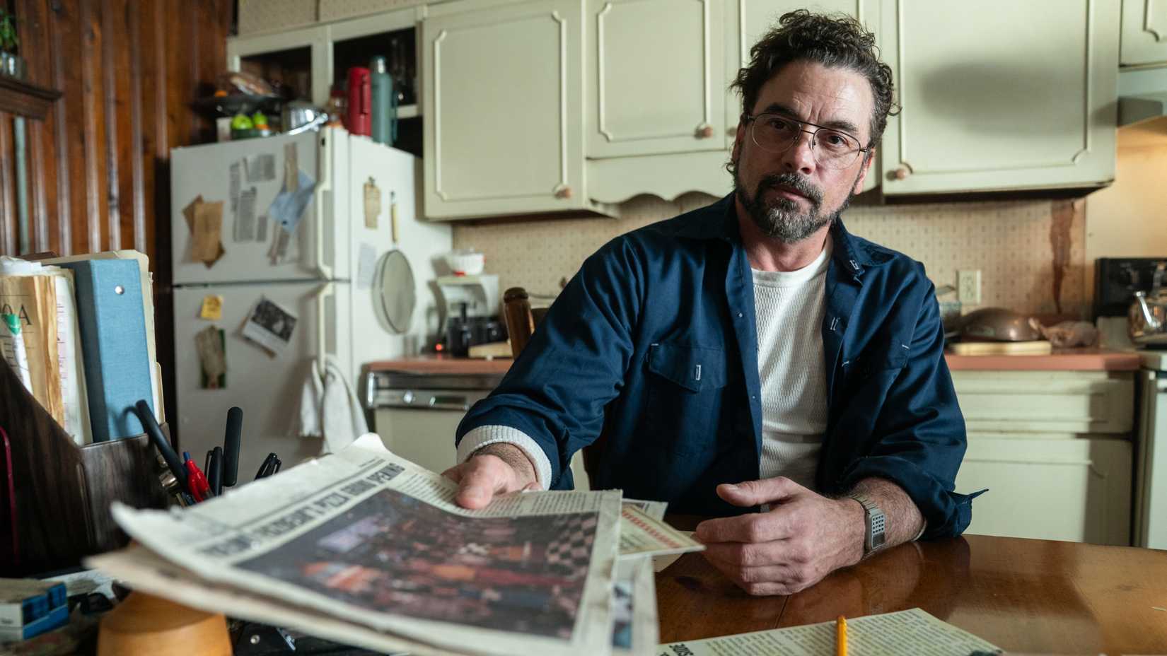 Skeet Ulrich sits at a table in a small kitchen, holding a newspaper out toward the camera, in Five Nights at Freddy's 2