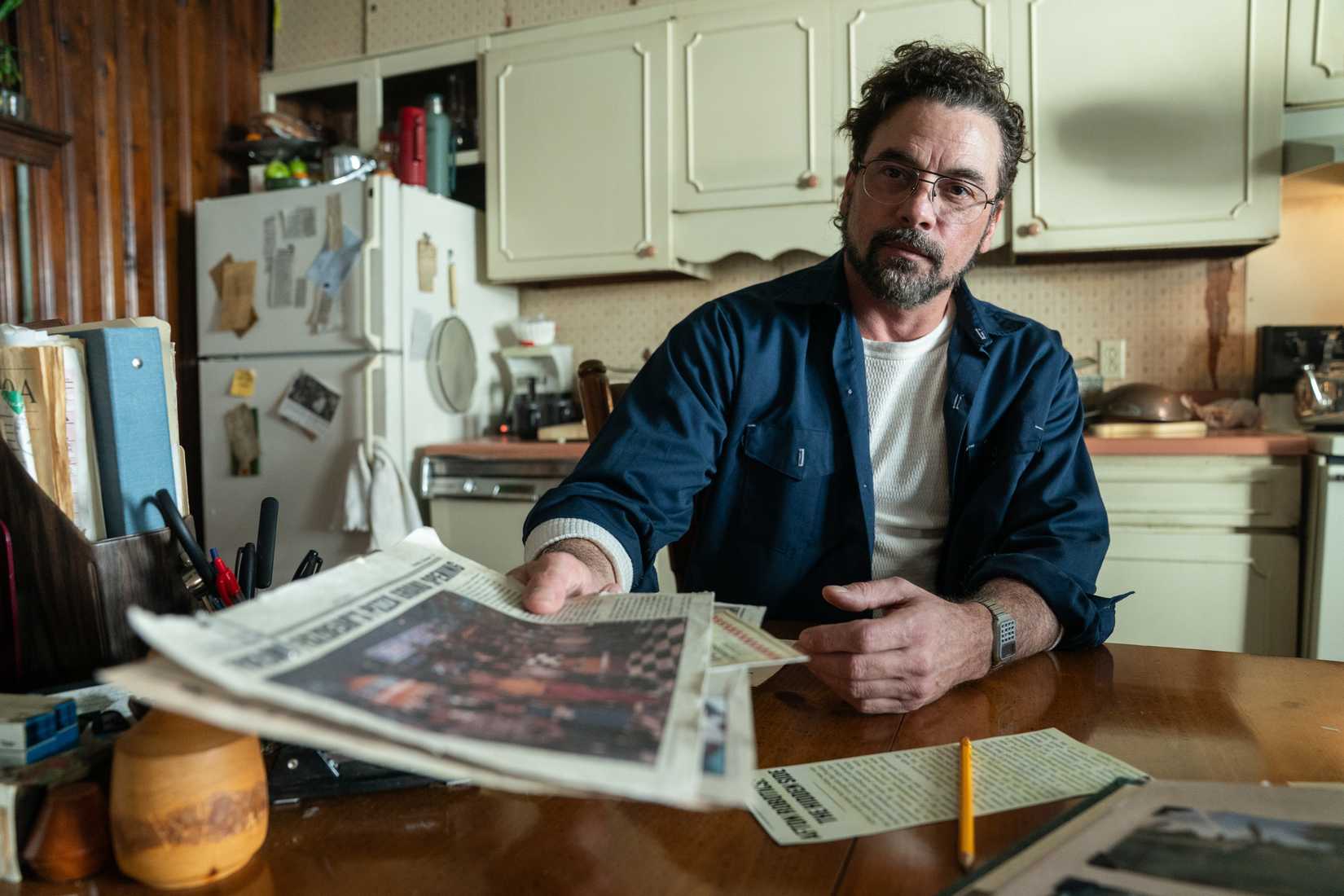 Skeet Ulrich sits at a table in a small kitchen, holding a newspaper out toward the camera, in Five Nights at Freddy's 2