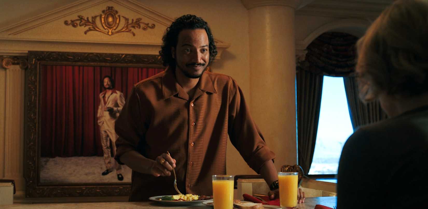 Koumba Diabaté (Samba Schutte) smiles warmly at Carol (Rhea Seehorn) over breakfast, with a painting of himself in the background, in Pluribus episode 6