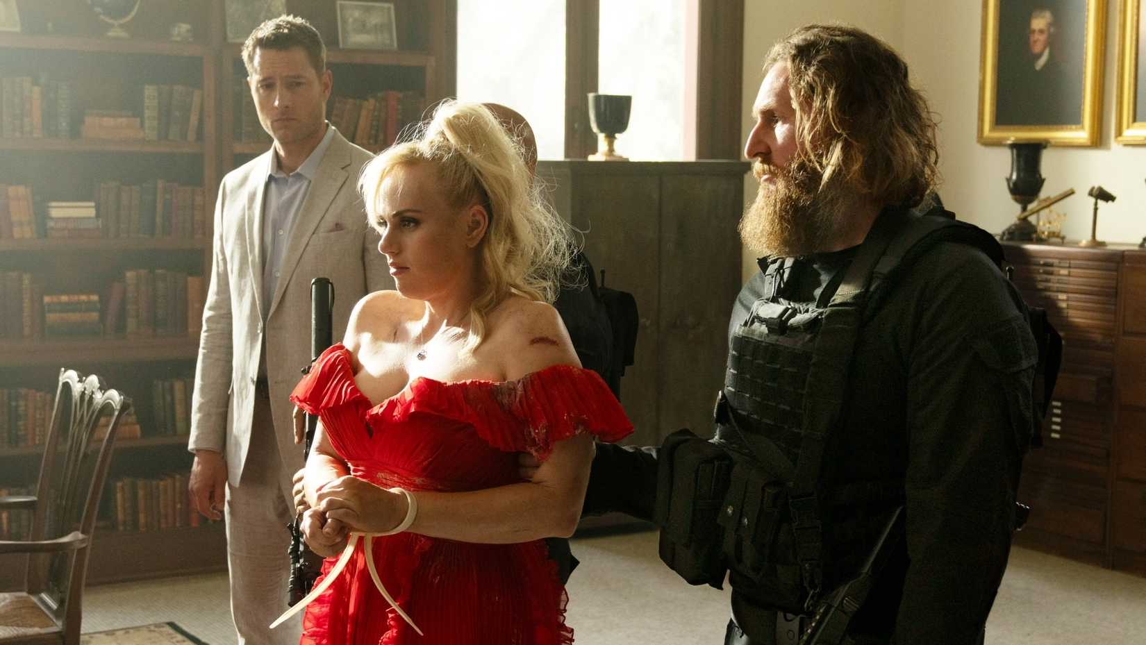 Rebel Wilson, wearing a bright red bridesmaid dress, is captured and bound by an armed man in a scene from the action-comedy Bride Hard.