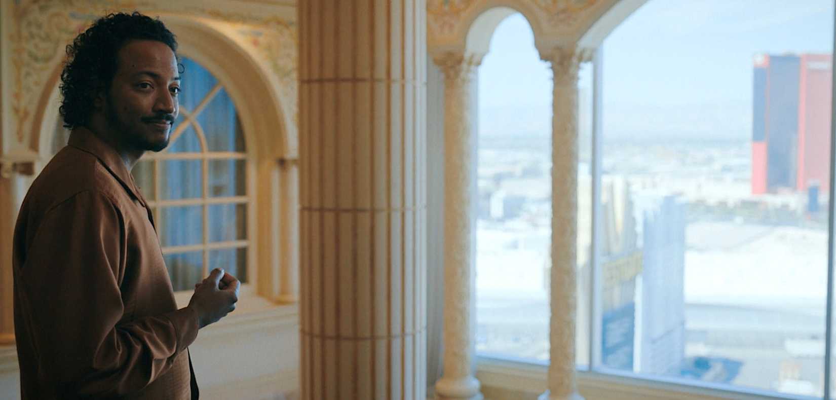 Koumba Diabaté (Samba Schutte) smiles warmly as he stands in front of a white marble column in Pluribus episode 6