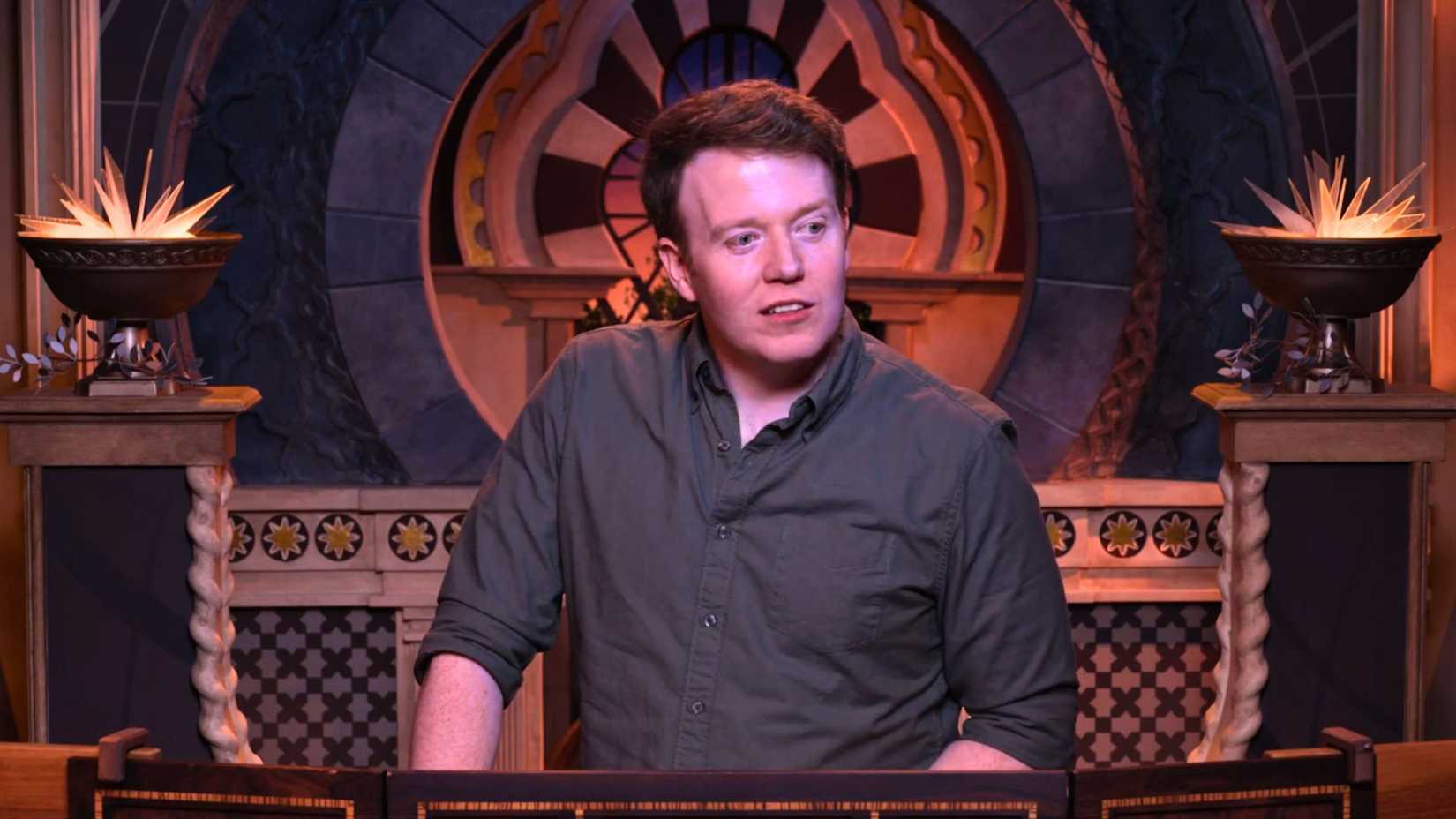 An image of Brennan Lee Mulligan from Critical Role Cooldown episode 9. He wears a grey shirt, and stands behind a DM screen.