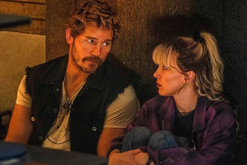 Chris Pratt and Millie Bobby Brown share rueful looks while sitting leaning against a wall in a scene from The Electric State