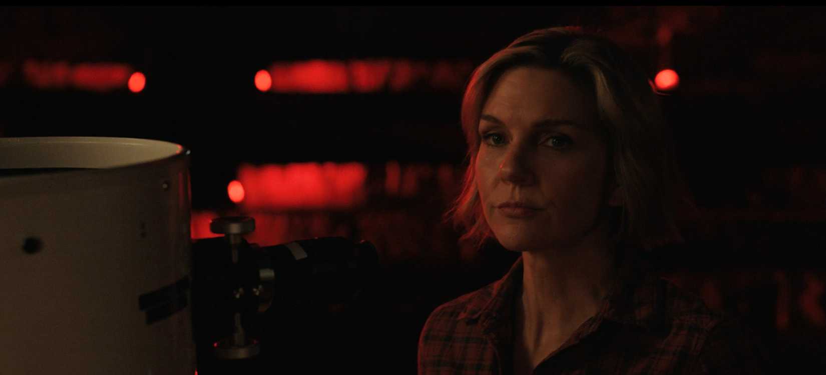 Carol (Rhea Seehorn) looks wry in a dark, red-tinted space, standing next to a telescope, in Pluribus