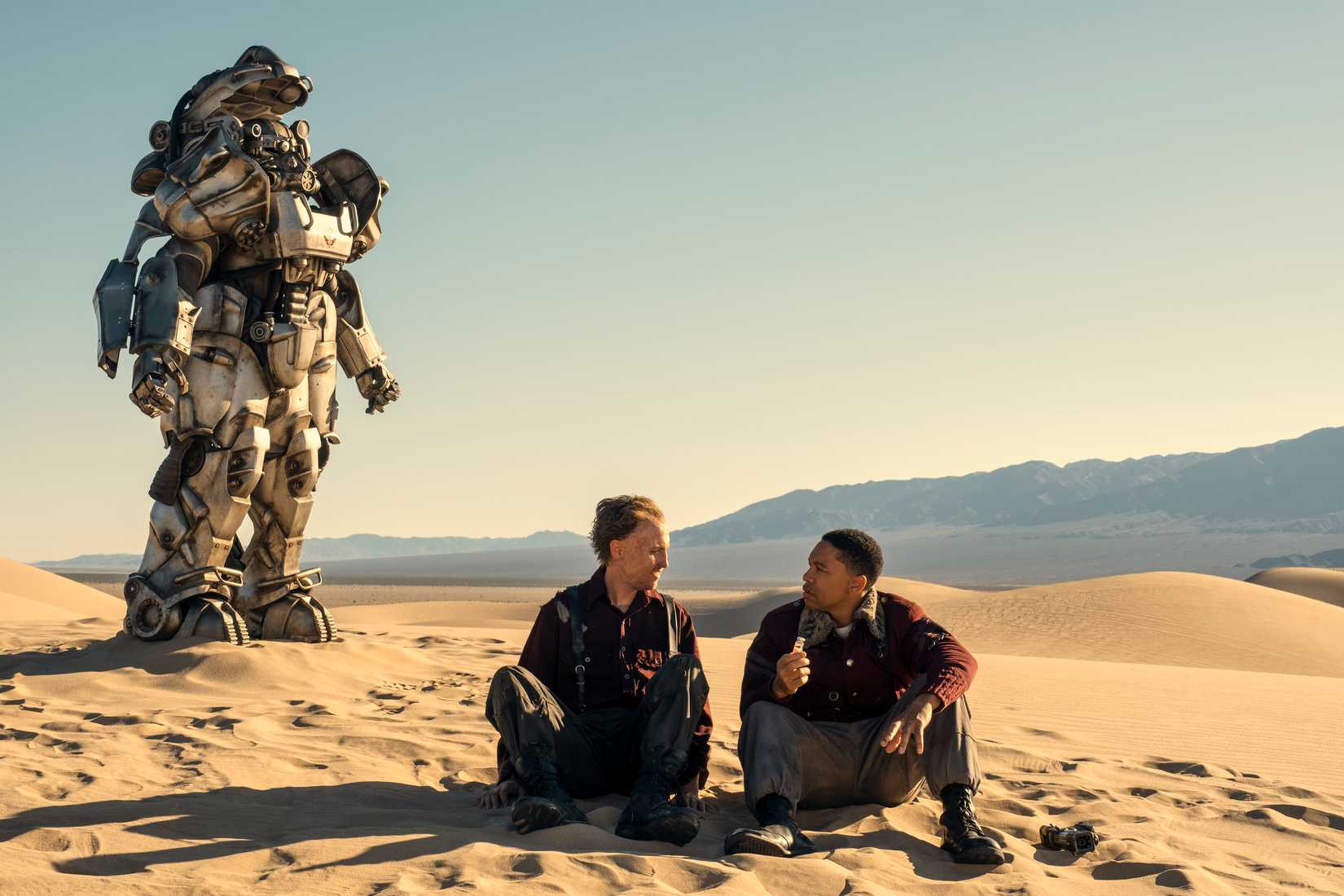 Johnny Pemberton (Thaddeus) and Aaron Moten (Maximus) sit in the sand with a suit of power armor behind them in Fallout season 2