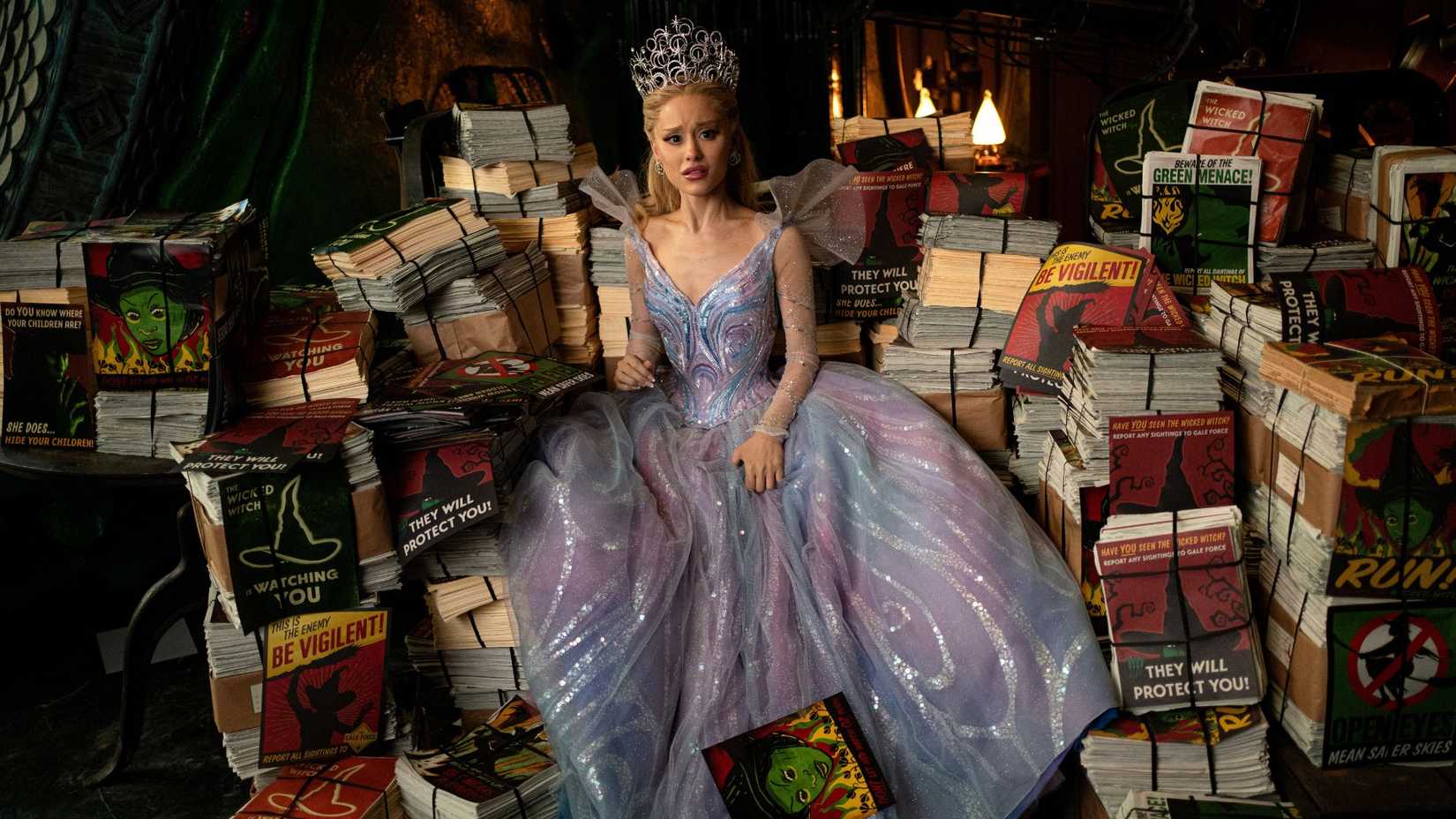 Glinda (Ariana Grande) sits on top a pile of different Wicked Witch of the West posters. She looks disgusted.