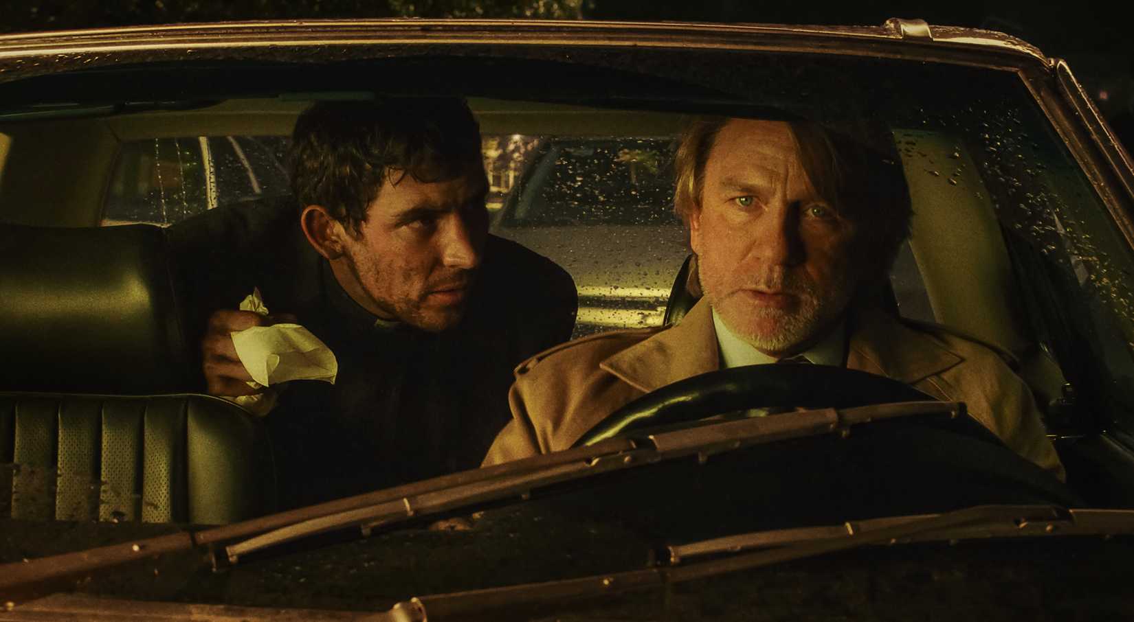 A blood-covered priest (Josh O'Connor) leans forward from the back seat of a car at night to speak to Benoit Blanc (Daniel Craig), who's in the driver's seat, in Wake Up Dead Man