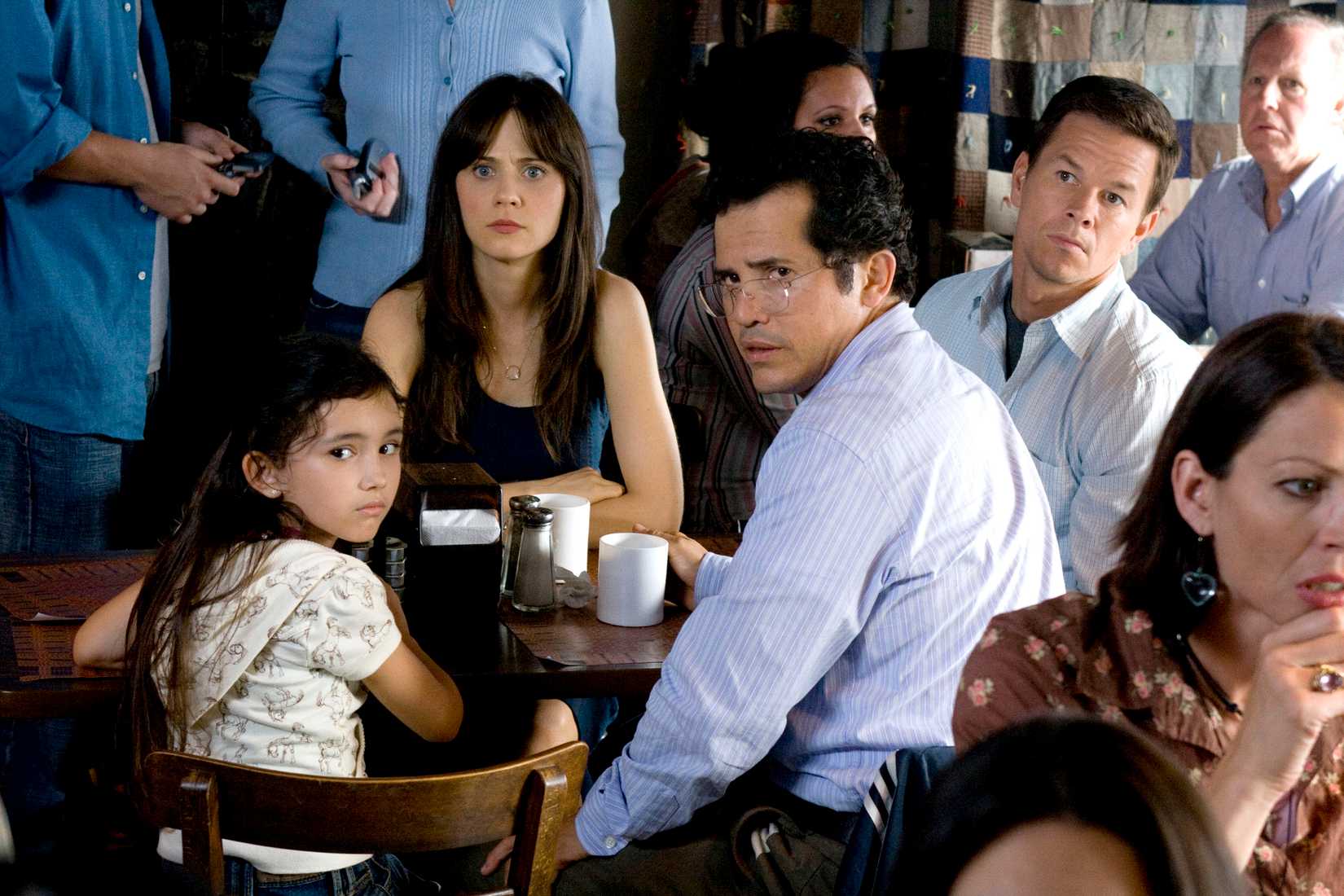 Ashlyn Sanchez, Zooey Deschanel, John Leguizamo, and Mark Wahlberg all sit together at a table in a crowded indoor space, staring at something off-screen, in The Happening.