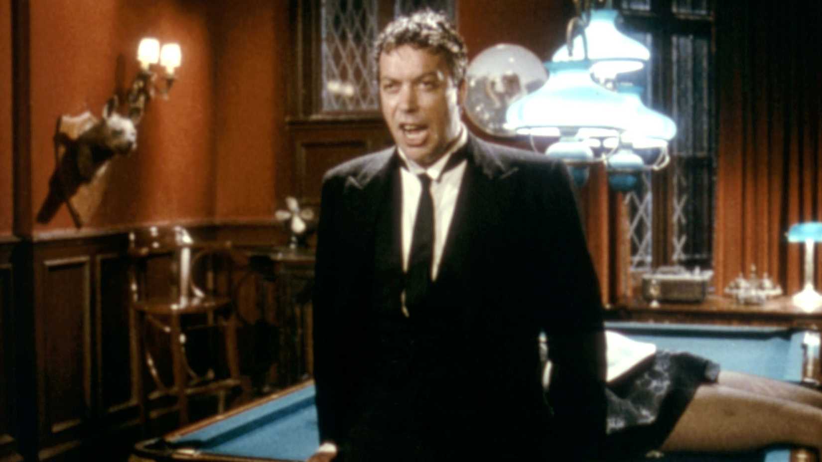 The butler Wadsworth (Tim Curry) explains the murder mystery while standing in front of a pool table with a corpse on it in Clue