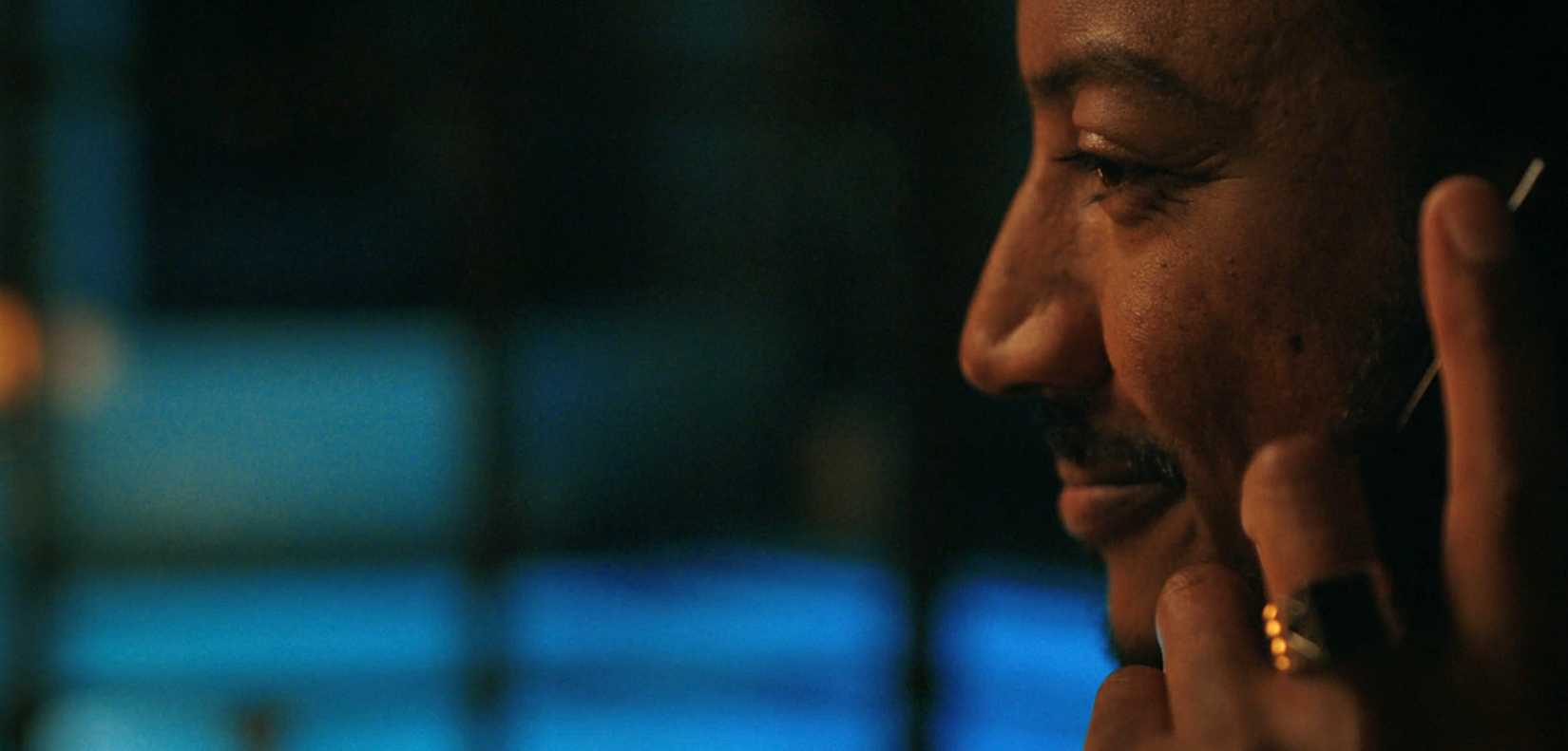 Koumba Diabaté (Samba Schutte) in extreme close-up, listening to someone on his cell phone and smiling in Pluribus episode 6