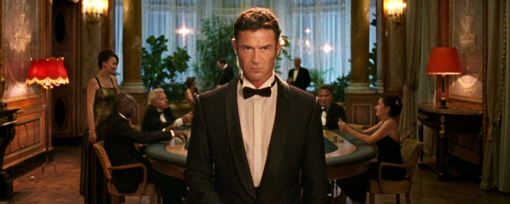 A stern-looking man in a tuxedo (Yannick Renier) stands in the middle of the screen, staring intently into the camera. Behind him, men in suits and women in cocktail dresses play cards around a casino table.