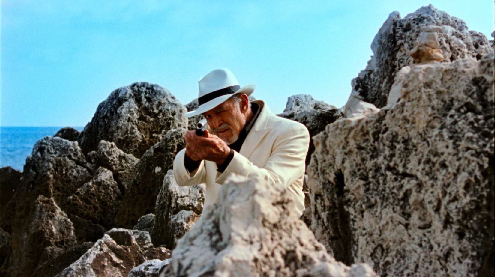 An older man in a white suit and fedora aims a rifle at the camera while standing amid huge porous rocks on the beach in Reflection in a Dead Diamond