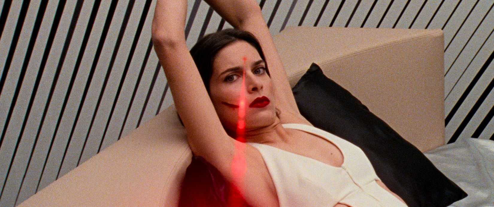 A woman in a white halter dress with her arms bound above her head lies back on a bed with a laser pointed at her forehead