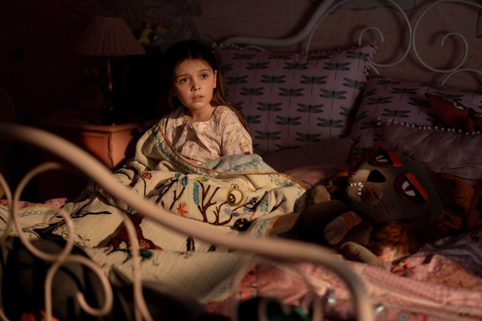 Sophie Sloan as Aurora, sitting on her bed in pajamas surrounded by toys and pillows.