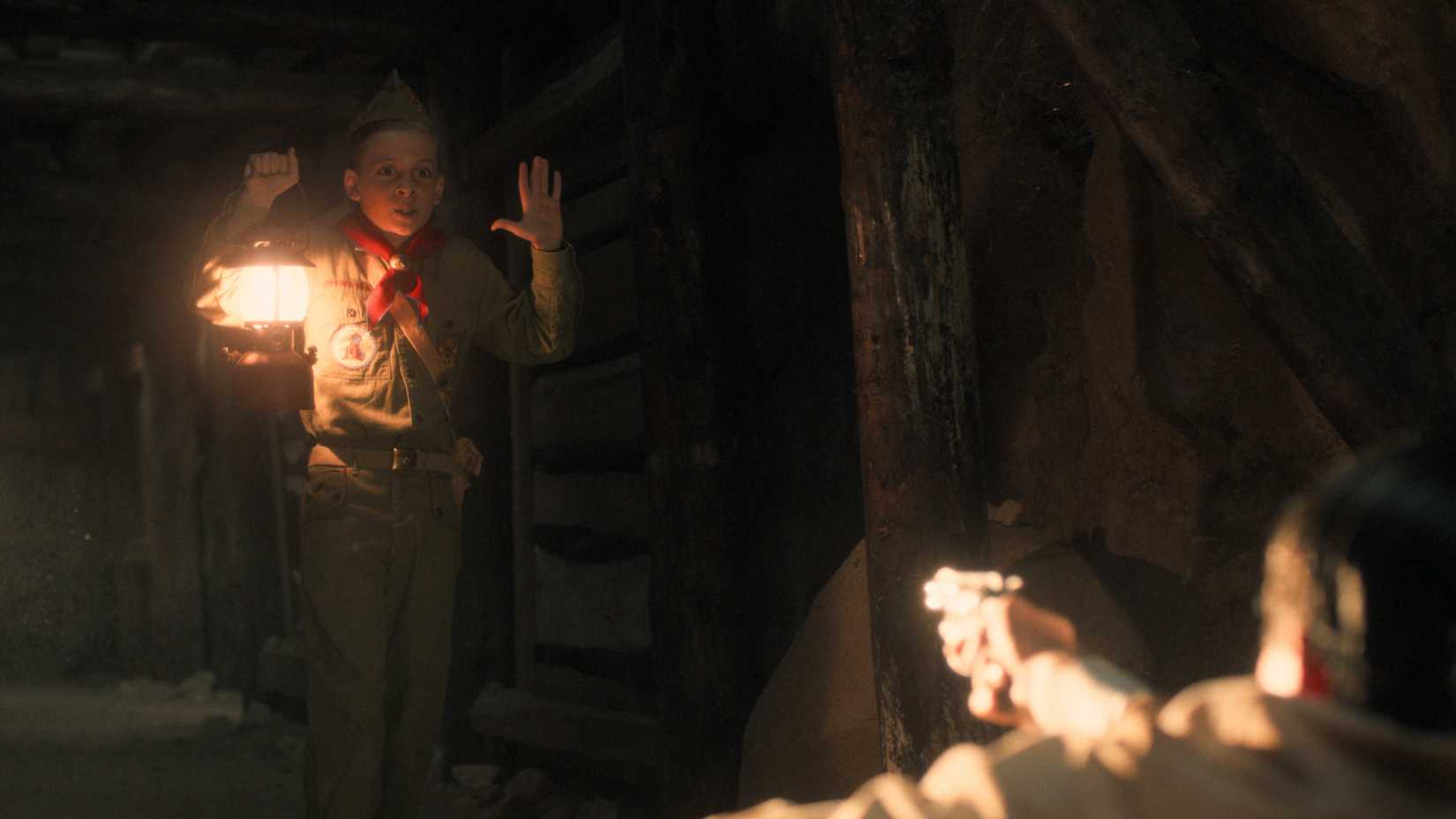 A young Henry Creel (Maksim Blatt) holds his hands up as a man a mineshaft in Nevada points a gun at him in Stranger Things season 5, episode 6