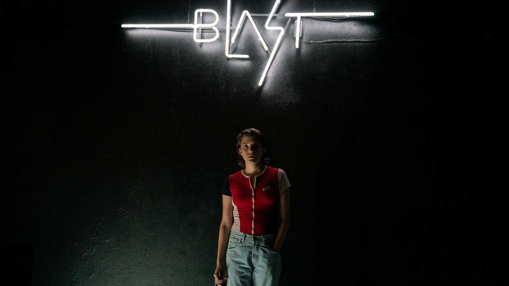 A young woman in a red shirt reclining beneath a neon sign that reads 'BLAST' at night