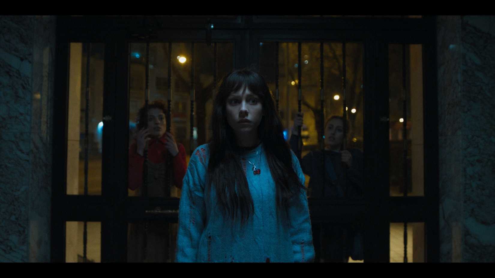 A nervous young woman in a blue jumper stands beneath the dim entrance of a block of flats