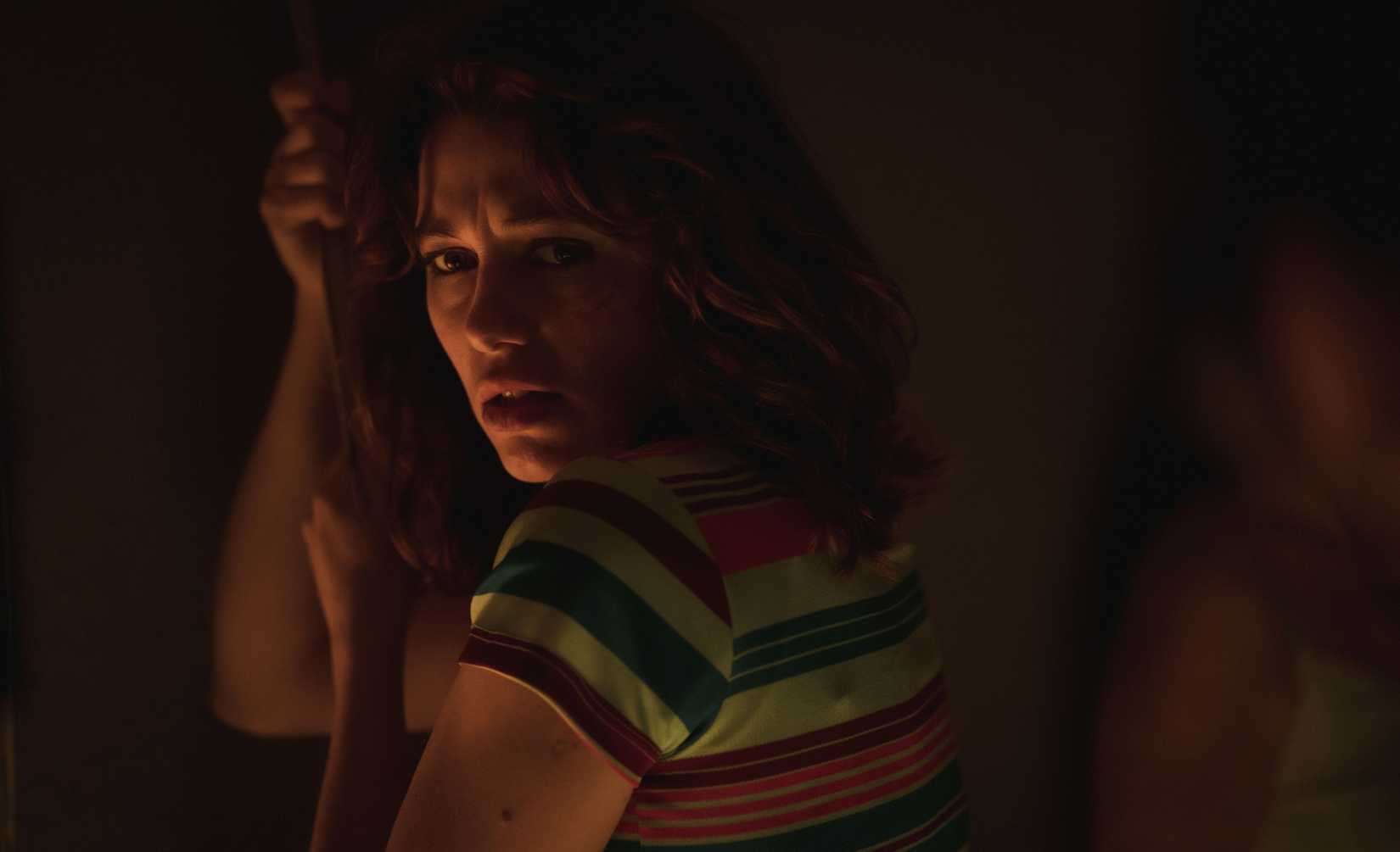 An image from The Wailing 2024. A woman with shoulder-length brown hair holds onto something dangling from the ceiling. She's in a dark room, and wears a black-and-white striped shirt. She looks a tad disturbed.