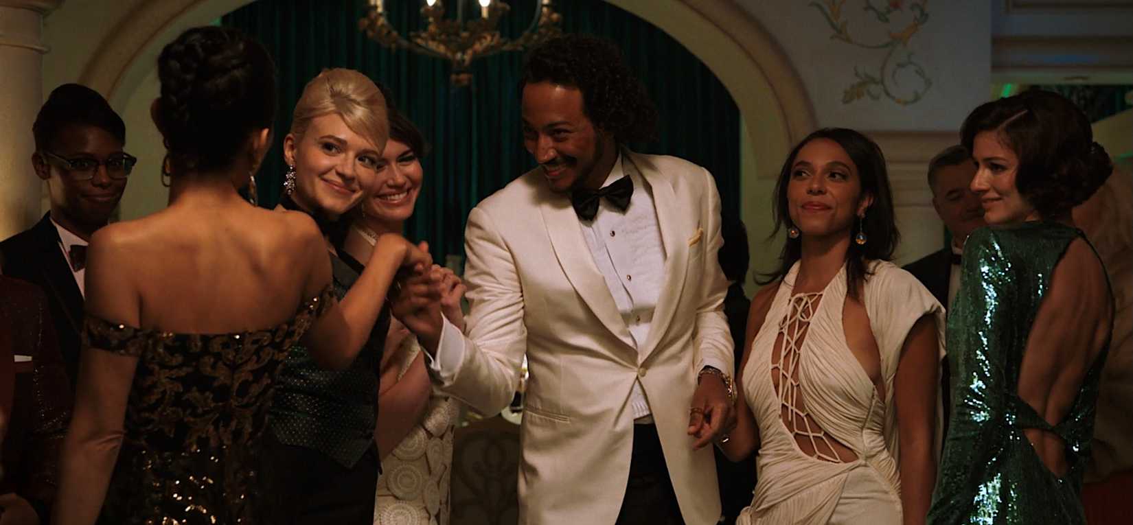 Diabaté (Samba Schutte), smiling and surrounded by beautiful women in formal party gowns, in Pluribus episode 6