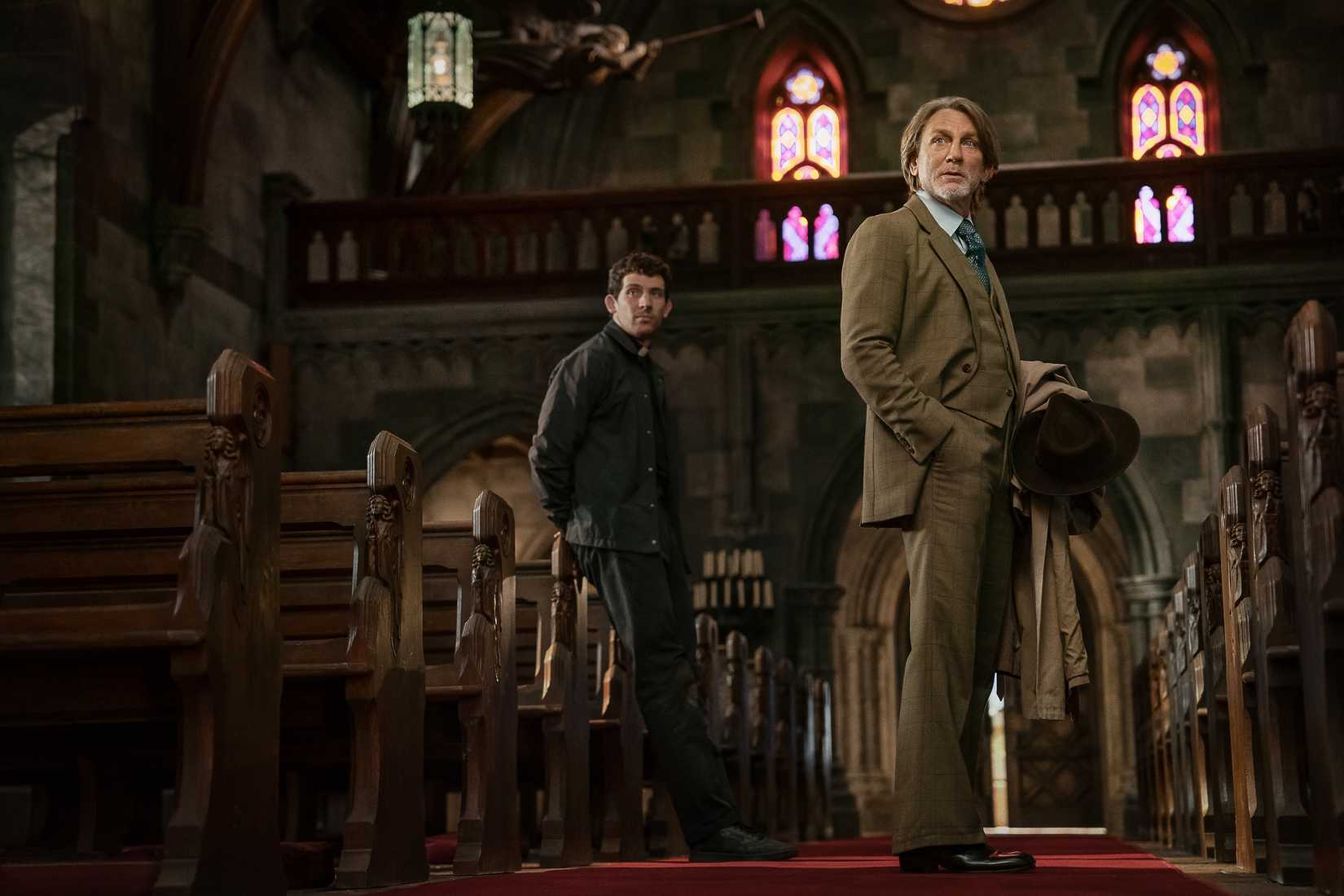 Two men (Josh O'Connor and Daniel Craig) stand in the aisle of a Catholic church, stained-glass windows above them, in Wake Up Dead Man