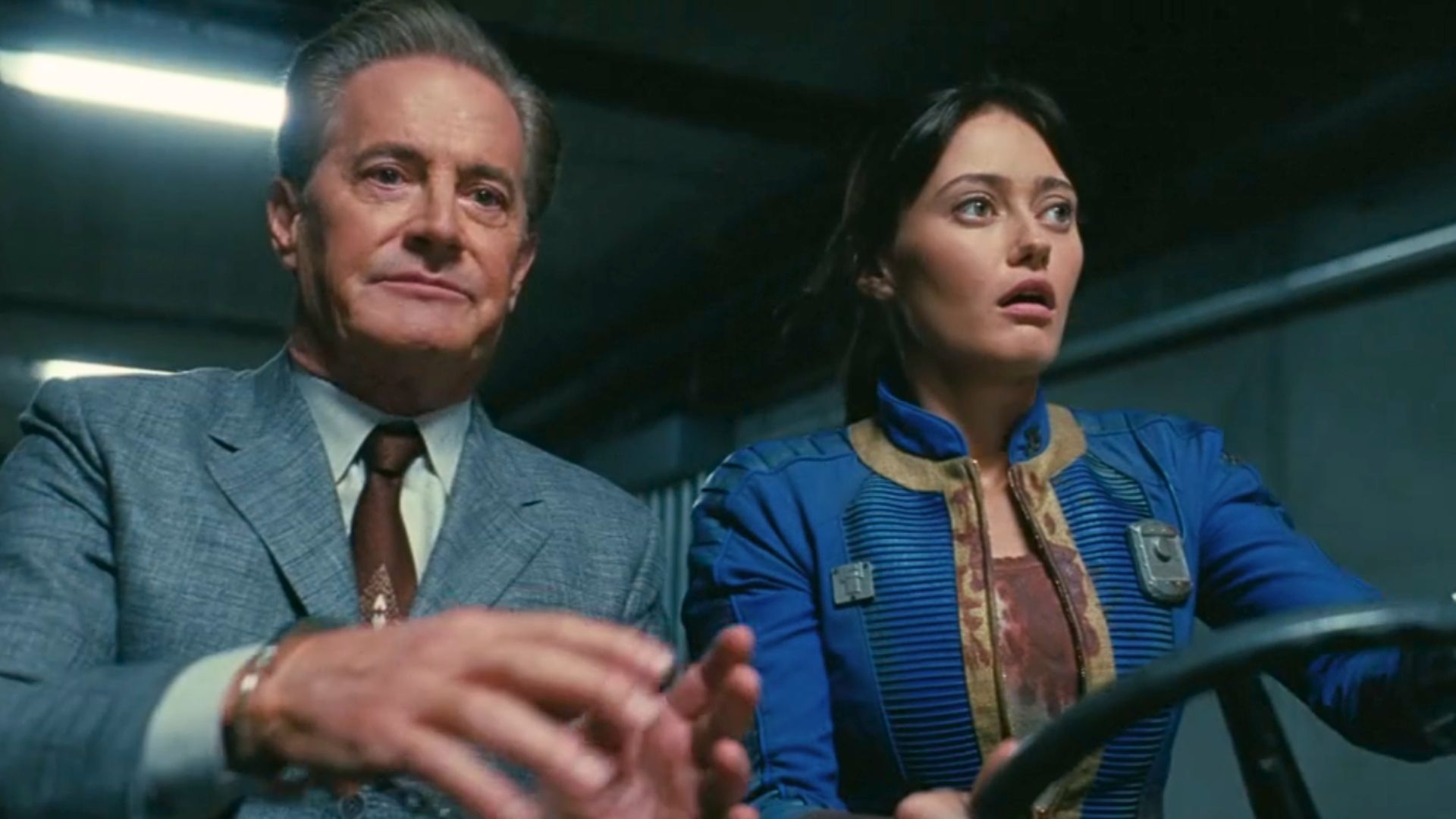 An image from Prime Video's Fallout season 2. It shows Hank, an older gentleman in a smart business suit, trying to take the wheel from Lucy, his daughter. She wears a blue vault suit.