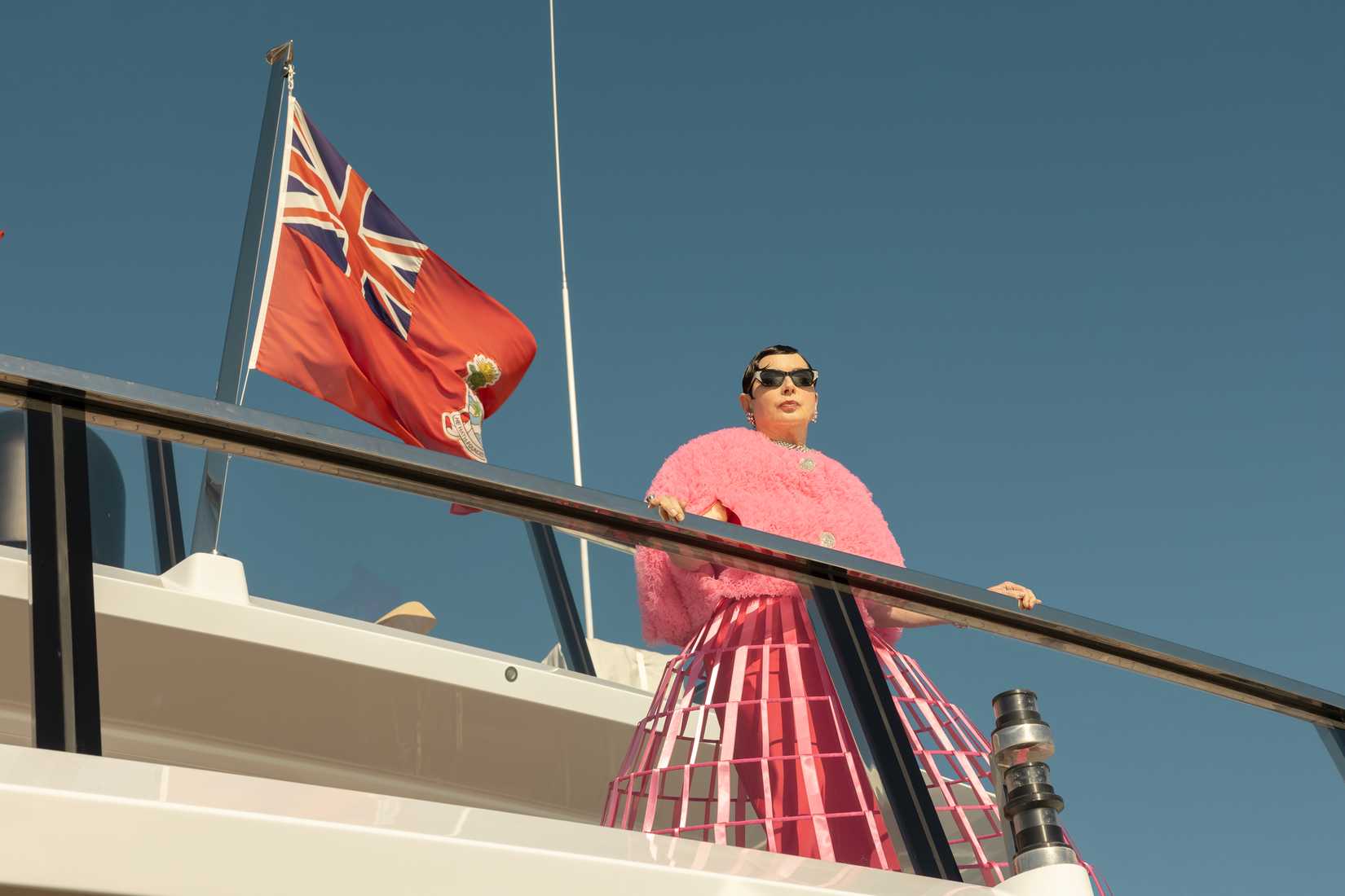 An image from The Beauty (2026). It shows a woman in a fluffy pink jacket, sunglasses, and a wirework ground-length skirt leaning against the railing of a yacht's upper deck.