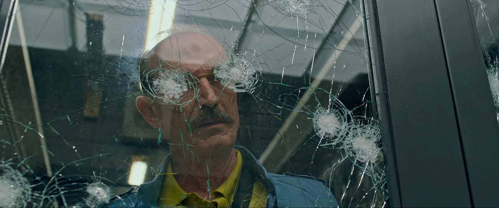 Toby Huss, playing a maniacal hitman, looks through a bullet-riddled window, with two of the bullet cracks lining up to cover his eyes, in a shot from Joe Carnahan's Copshop (2021)