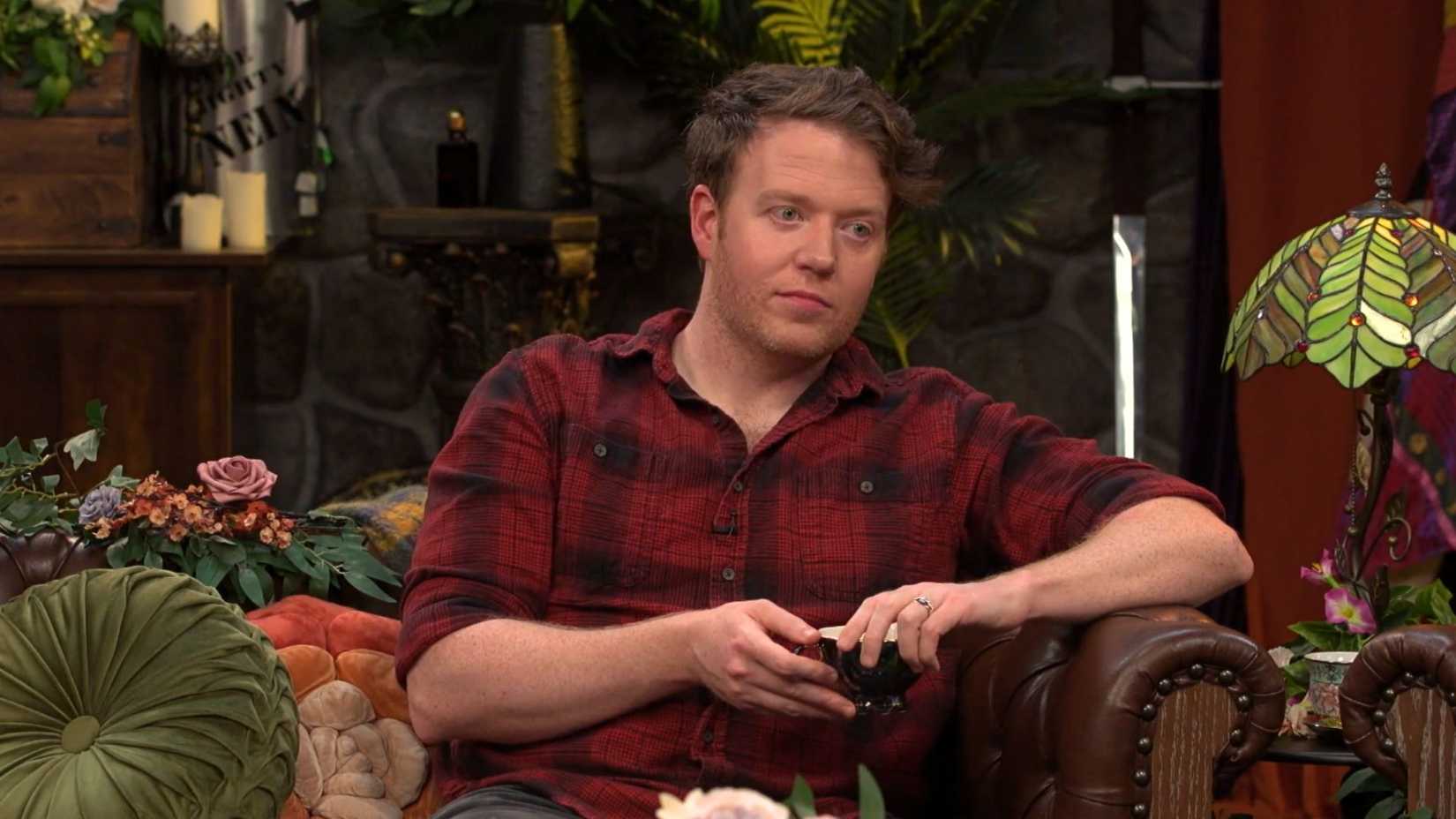Brennan Lee Mulligan relaxes on the Critical Role set in a flannel shirt, holding a teacup during a Tale Gate discussion.