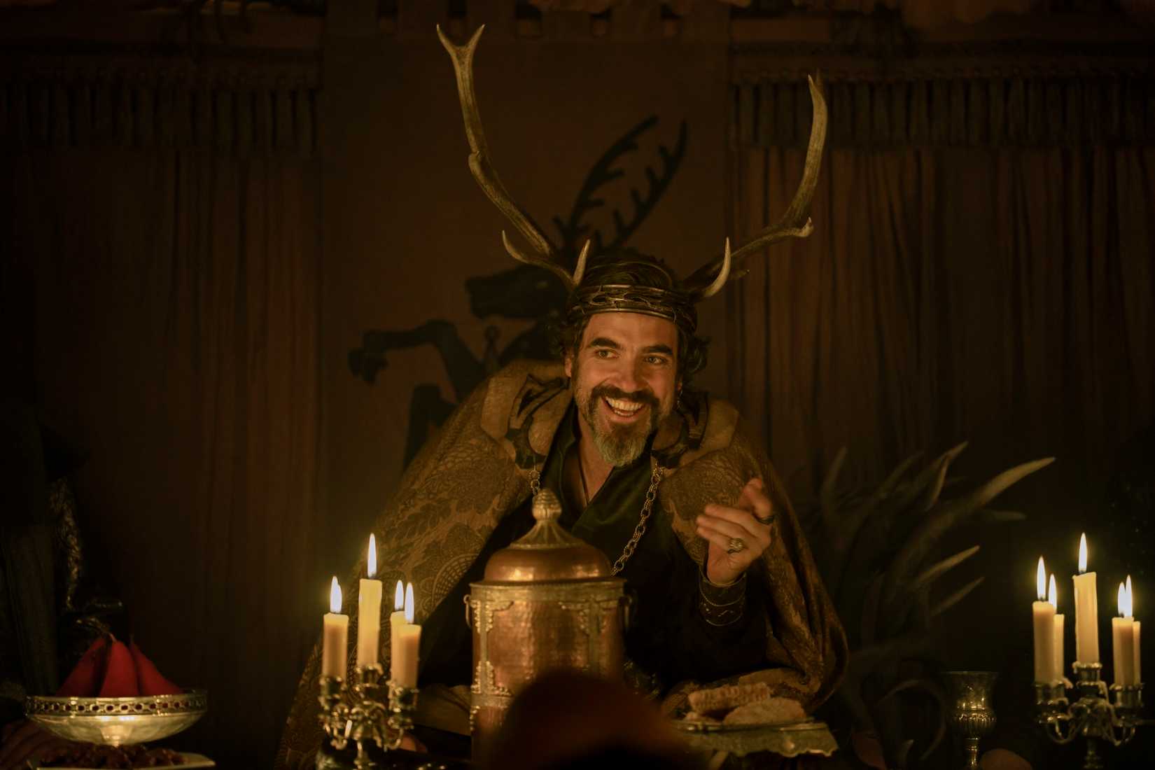 Ser Lionel Baratheon (Daniel Ings), in a furry leather cloak and a headdress with spreading deer antlers, sits at a candle-covered table and grins in A Knight of the Seven Kingdoms