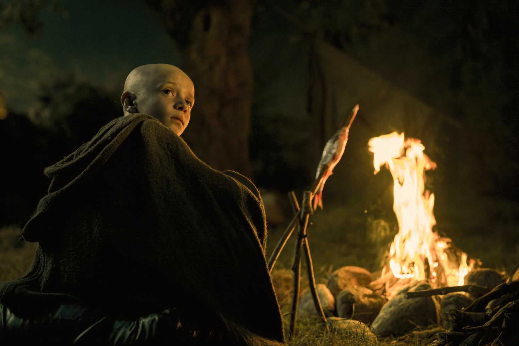 Egg (Dexter Sol Ansell), um menino pequeno e careca com uma grande capa marrom, está sentado perto de uma fogueira à noite no prelúdio de Game of Thrones, Um Cavaleiro dos Sete Reinos. Foto: Steffan Hill/HBO Max
