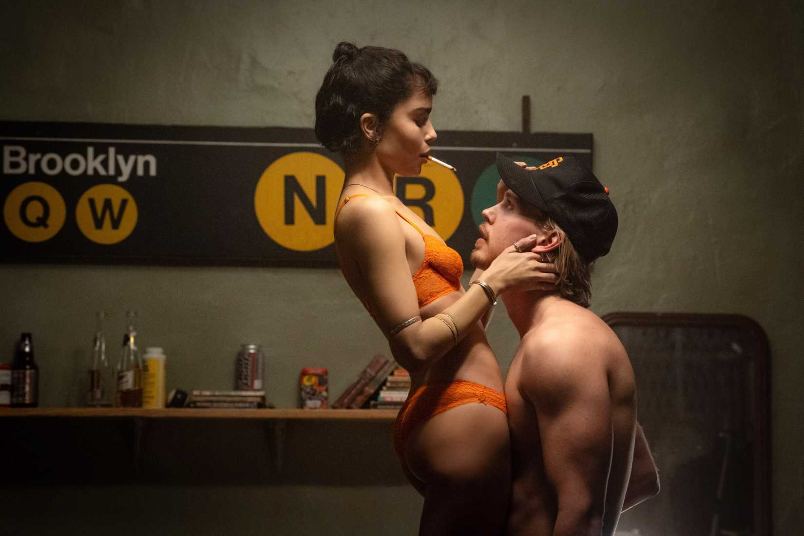 Zoë Kravitz in an orange underwear smokes and sits on Austin BUtler's lap in front of a New York subway sign in Caught Stealing