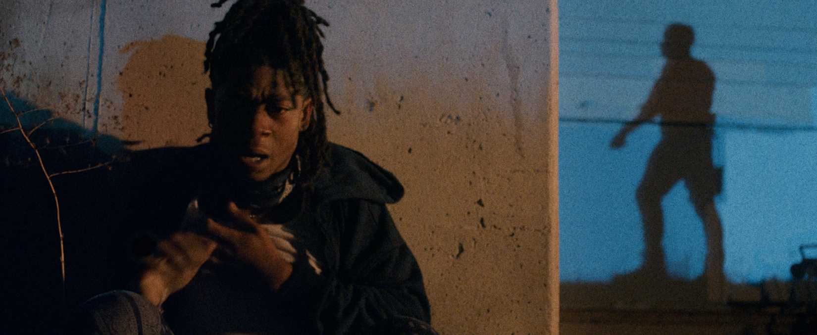 A thin Black man with dreadlocks cringes behind a wall at night as a silhouetted armed police officer hunts him in Night Patrol