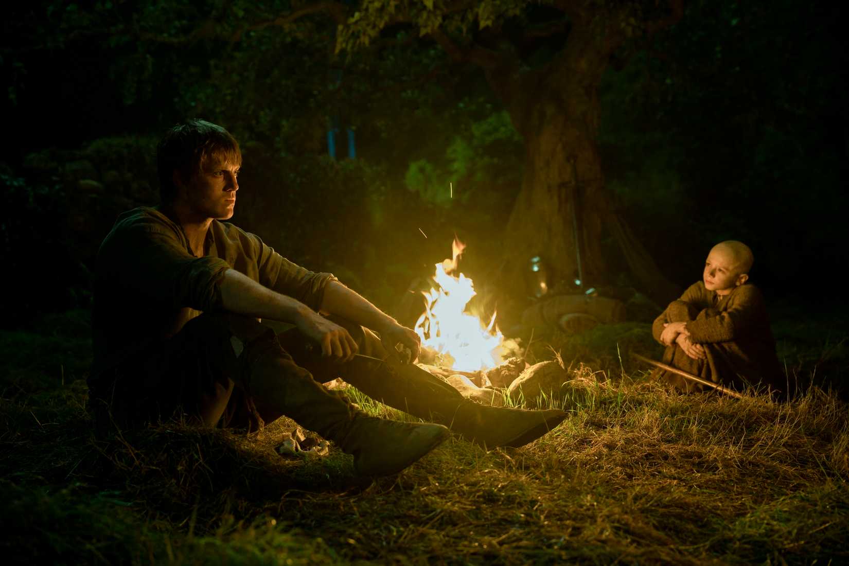 Dunk (Peter Claffey), a tall man in medieval clothing, sits pensively by a fire at night, across from Egg, a bald-headed child, in the Game of Thrones prequel A Knight of the Seven Kingdoms