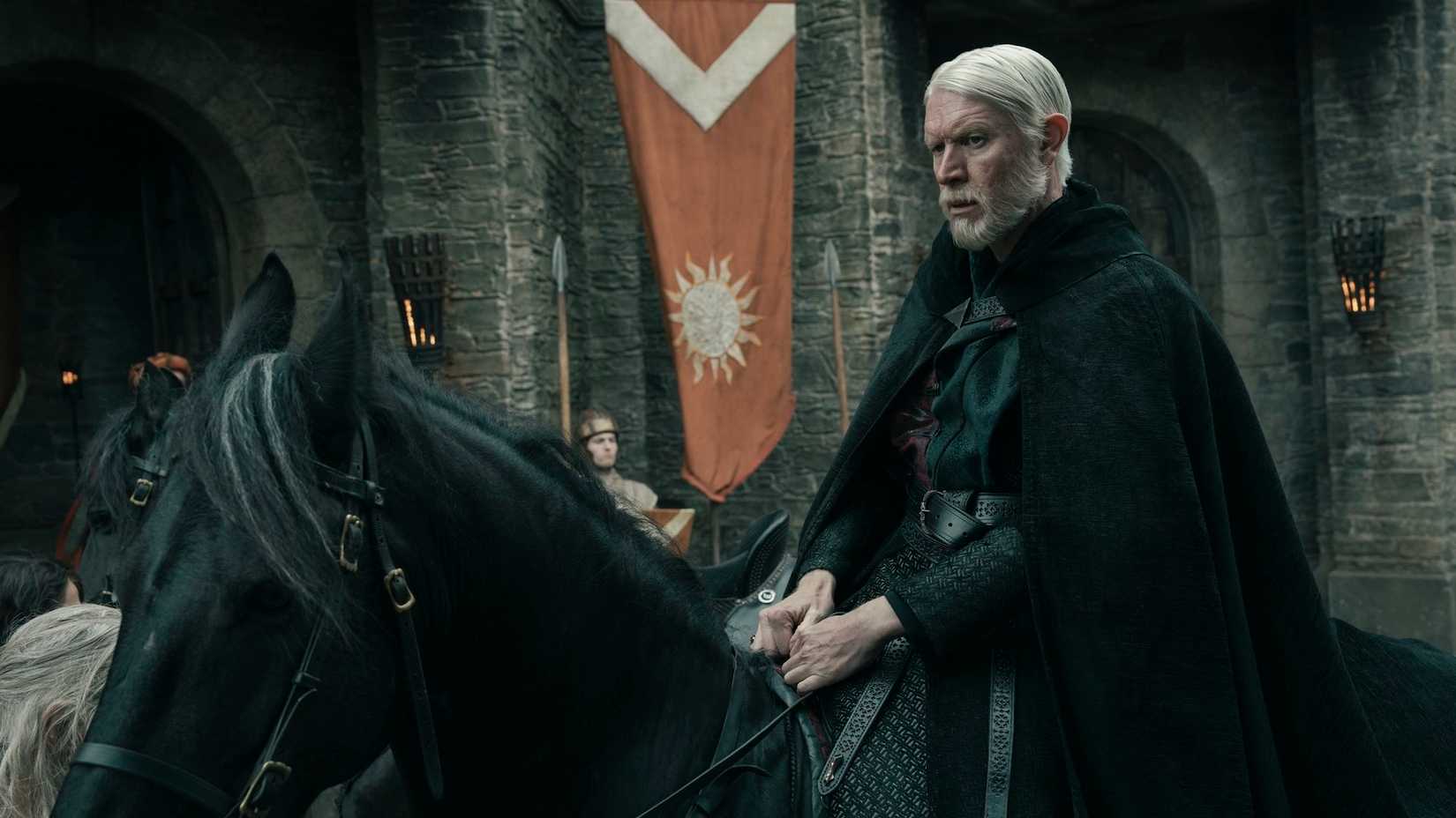 Maekar Targaryen (Sam Spruell) is dressed all in black sitting on top of a black horse in front of a castle in A Knight of the Seven Kingdoms