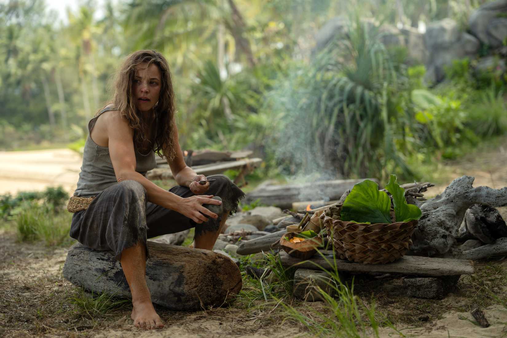 Linda (Rachel McAdams), a woman in filthy, bedraggled clothing, sits on a log by a hand-built fire-pit among scavenged fruits and a hand-woven basket in Sam Raimi's Send Help