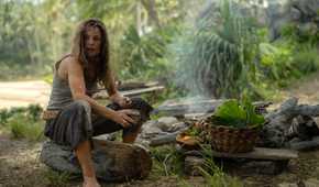 Linda (Rachel McAdams), a woman in filthy, bedraggled clothing, sits on a log by a hand-built fire-pit among scavenged fruits and a hand-woven basket in Sam Raimi's Send Help