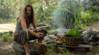 Linda (Rachel McAdams), a woman in filthy, bedraggled clothing, sits on a log by a hand-built fire-pit among scavenged fruits and a hand-woven basket in Sam Raimi's Send Help