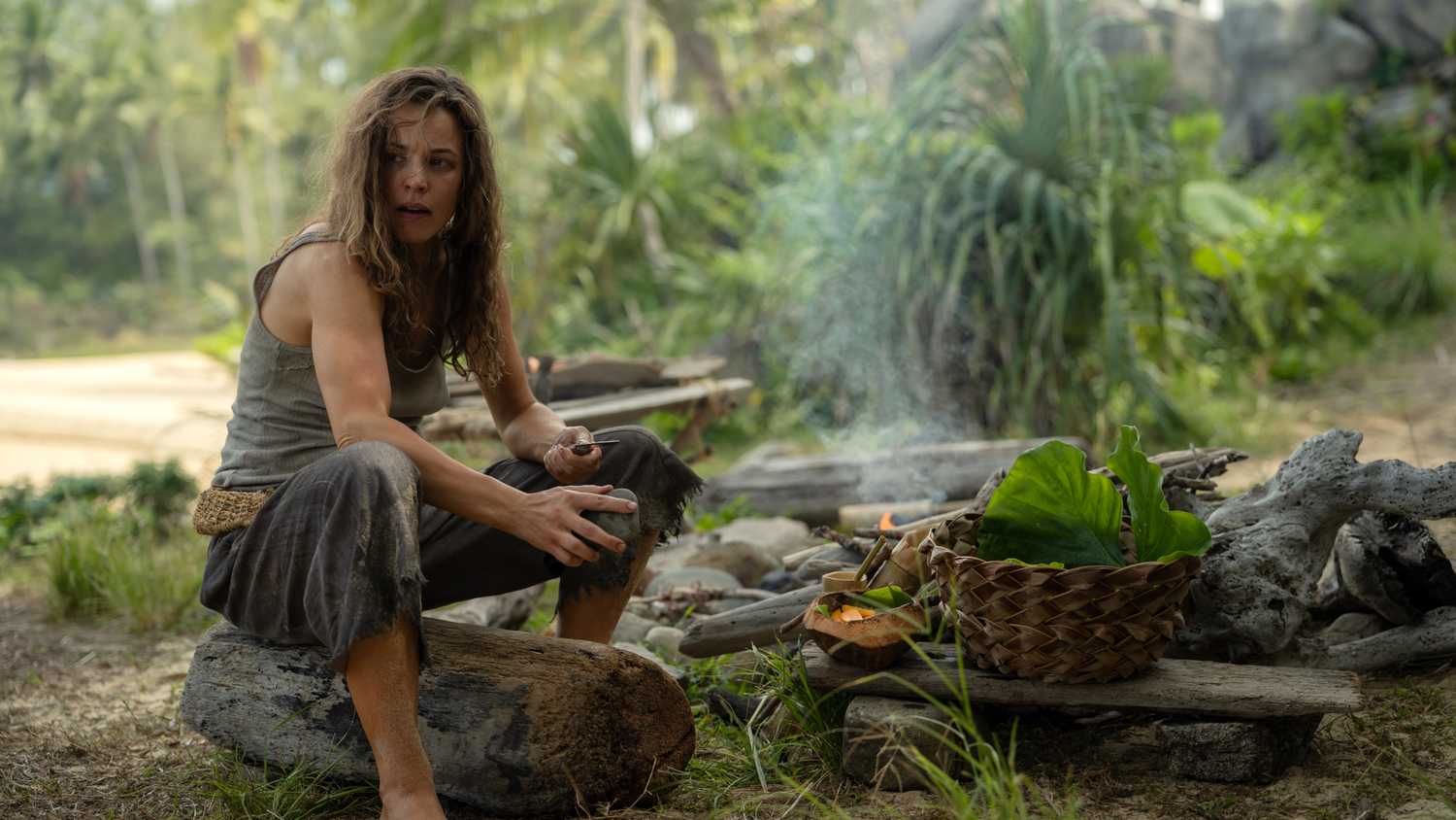 Linda (Rachel McAdams), a woman in filthy, bedraggled clothing, sits on a log by a hand-built fire-pit among scavenged fruits and a hand-woven basket in Sam Raimi's Send Help