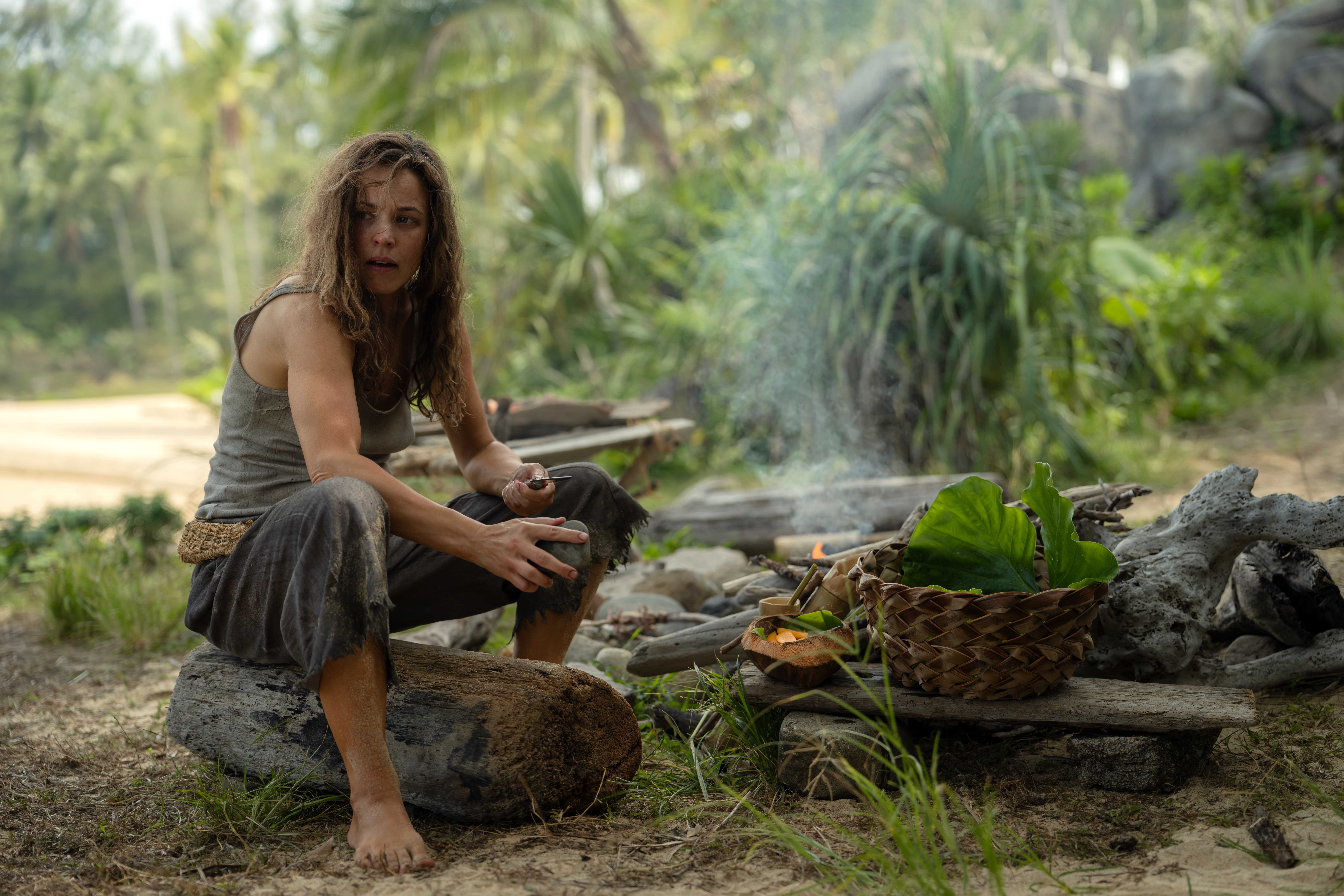 Linda (Rachel McAdams), a woman in filthy, bedraggled clothing, sits on a log by a hand-built fire-pit among scavenged fruits and a hand-woven basket in Sam Raimi's Send Help