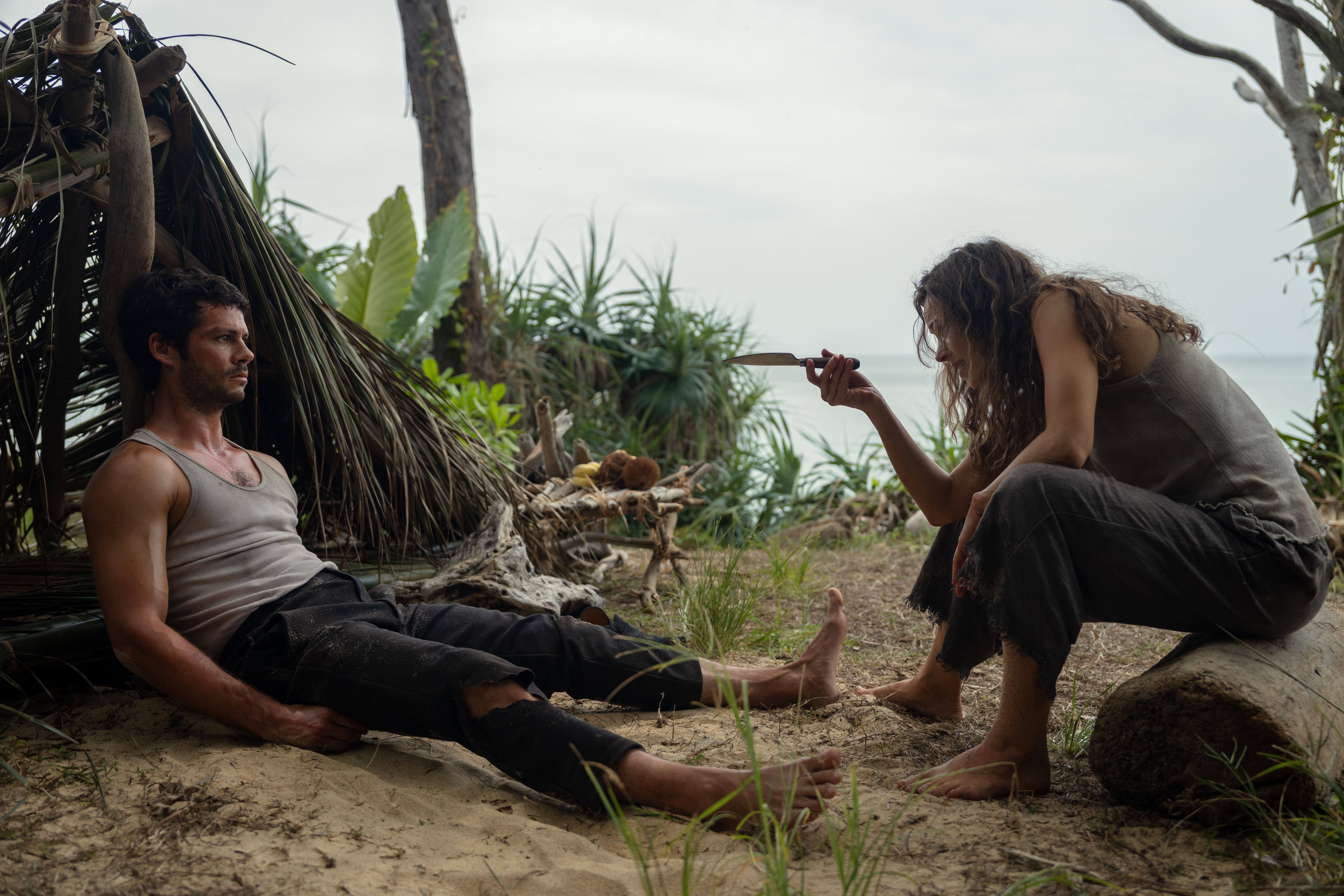 Brad (Dylan O'Brien), a barefoot, bedraggled man in ripped pants and a filthy sleeveless T-shirt, sprawls on sand in front of a shelter made out of fronds and leaves, as Linda (Rachel McAdams), a similarly grimy woman, gestures at him with a knife in Sam Raimi's Send Help